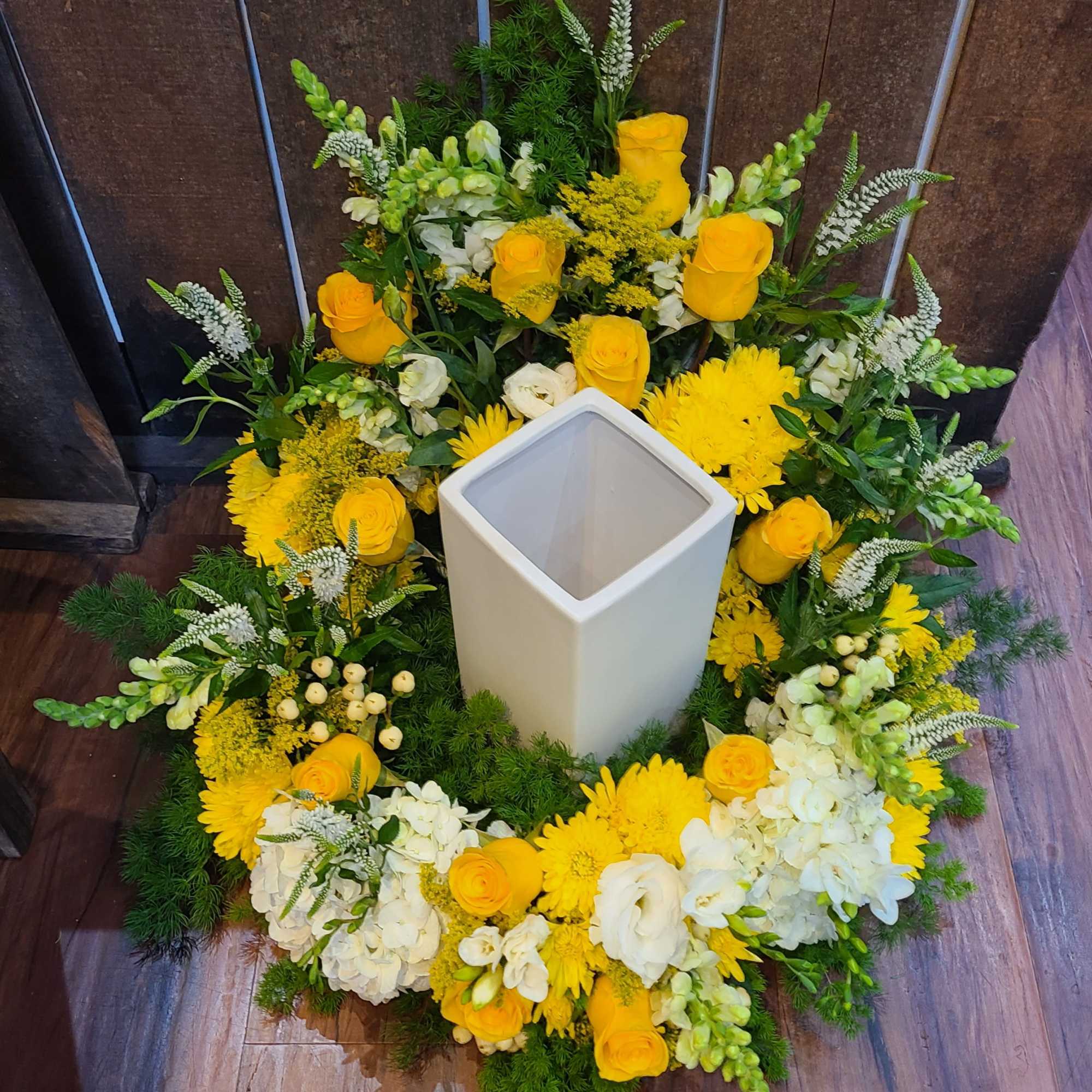 Circular yellow and white floral ring with roses and mums around a tall white square vase.