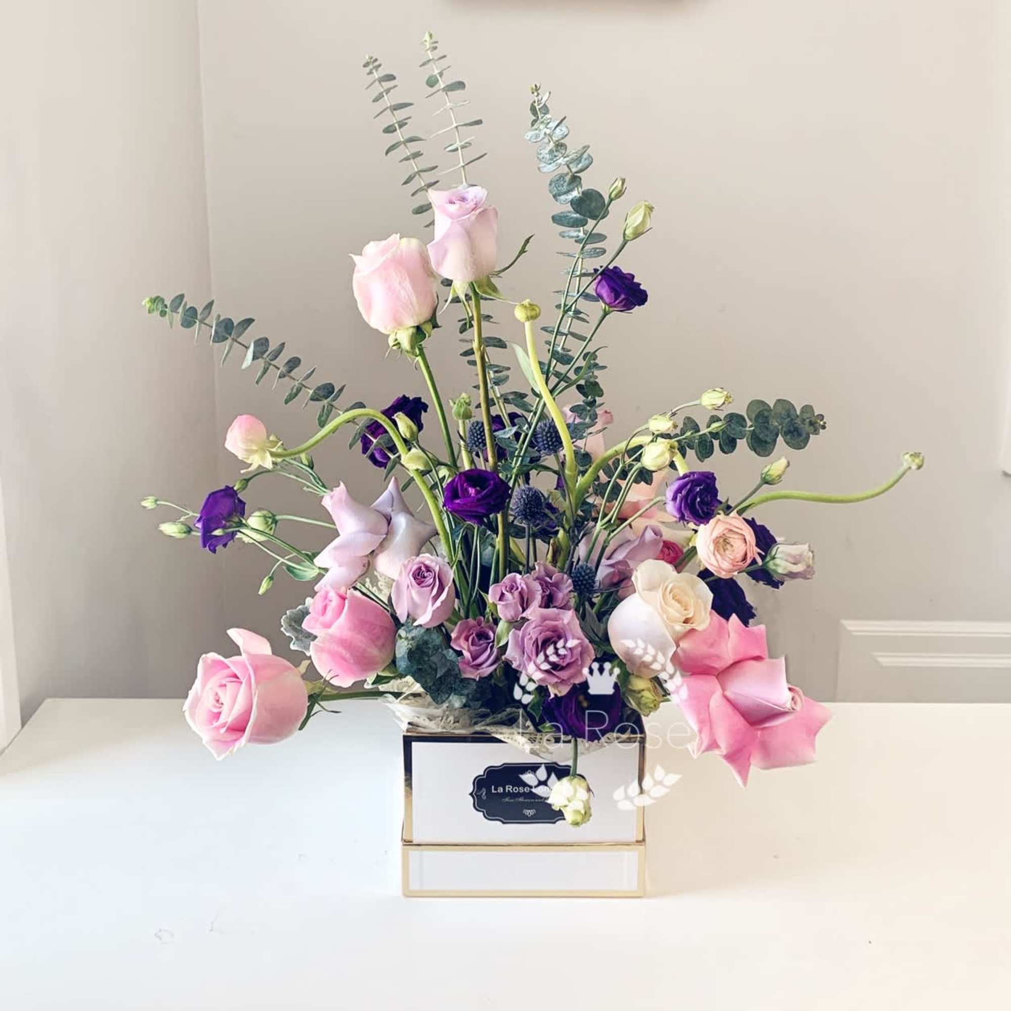 Pink and purple roses arranged in a white box with tall eucalyptus stems.
