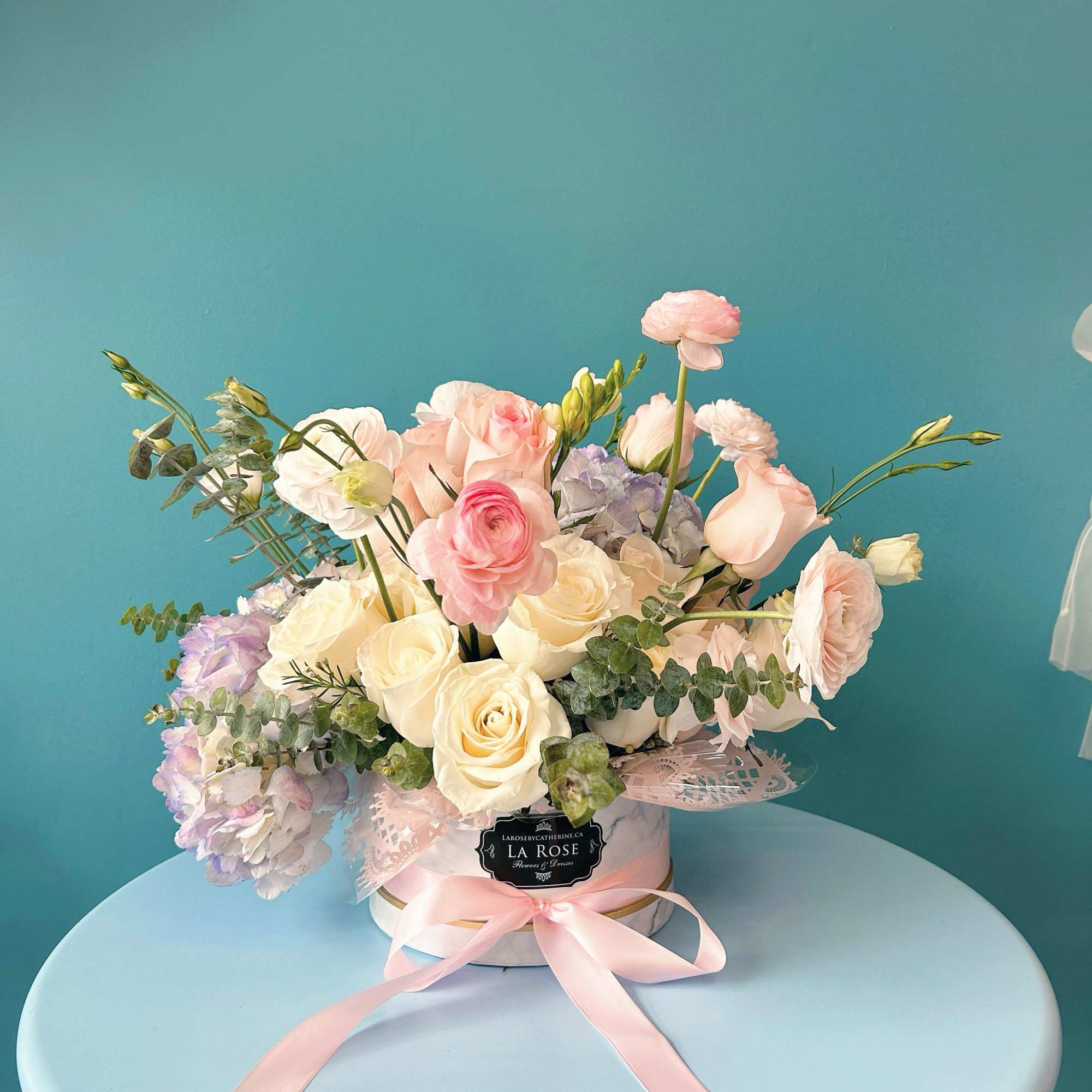 Pastel bouquet of roses, ranunculus, and hydrangeas in a round box with a pink ribbon