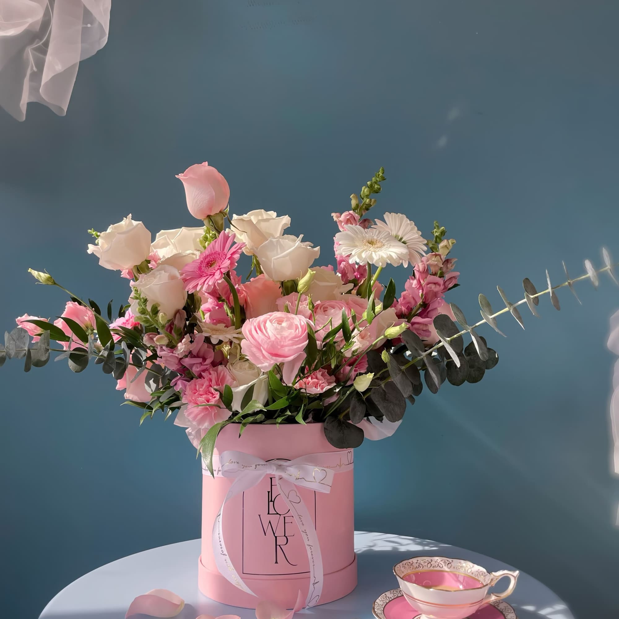 Pink and white flower arrangement in a pink box with a teacup and saucer