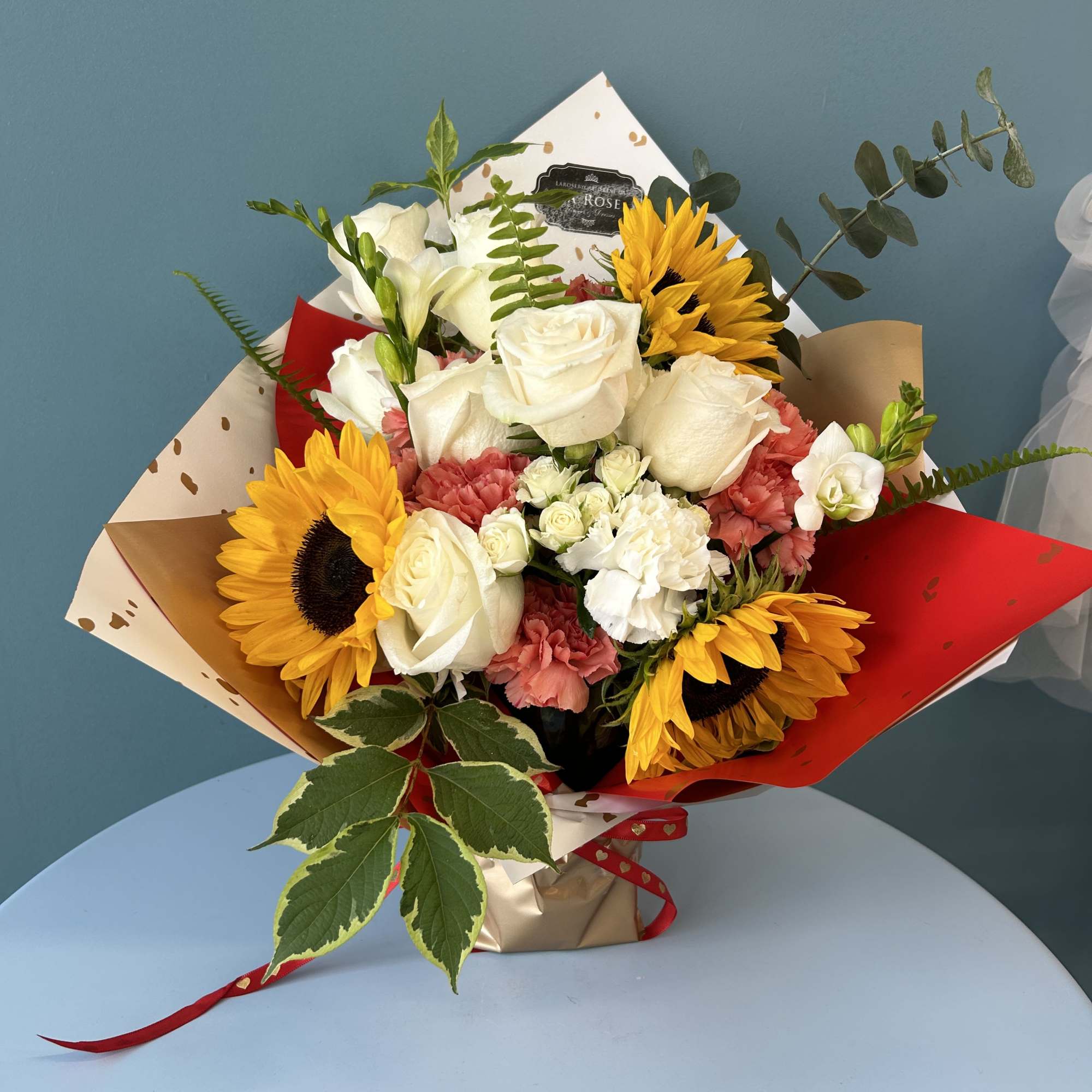 Bouquet of white roses, sunflowers, and pink carnations wrapped in red and beige paper