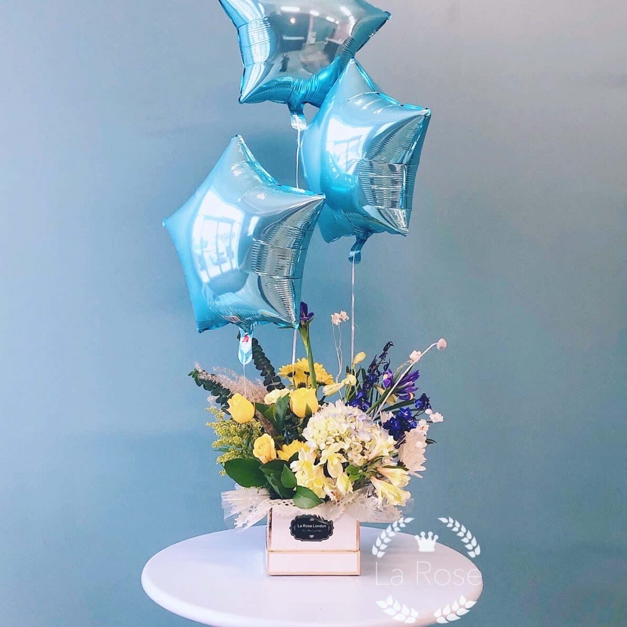 Yellow and white flower arrangement with blue star balloons in a box