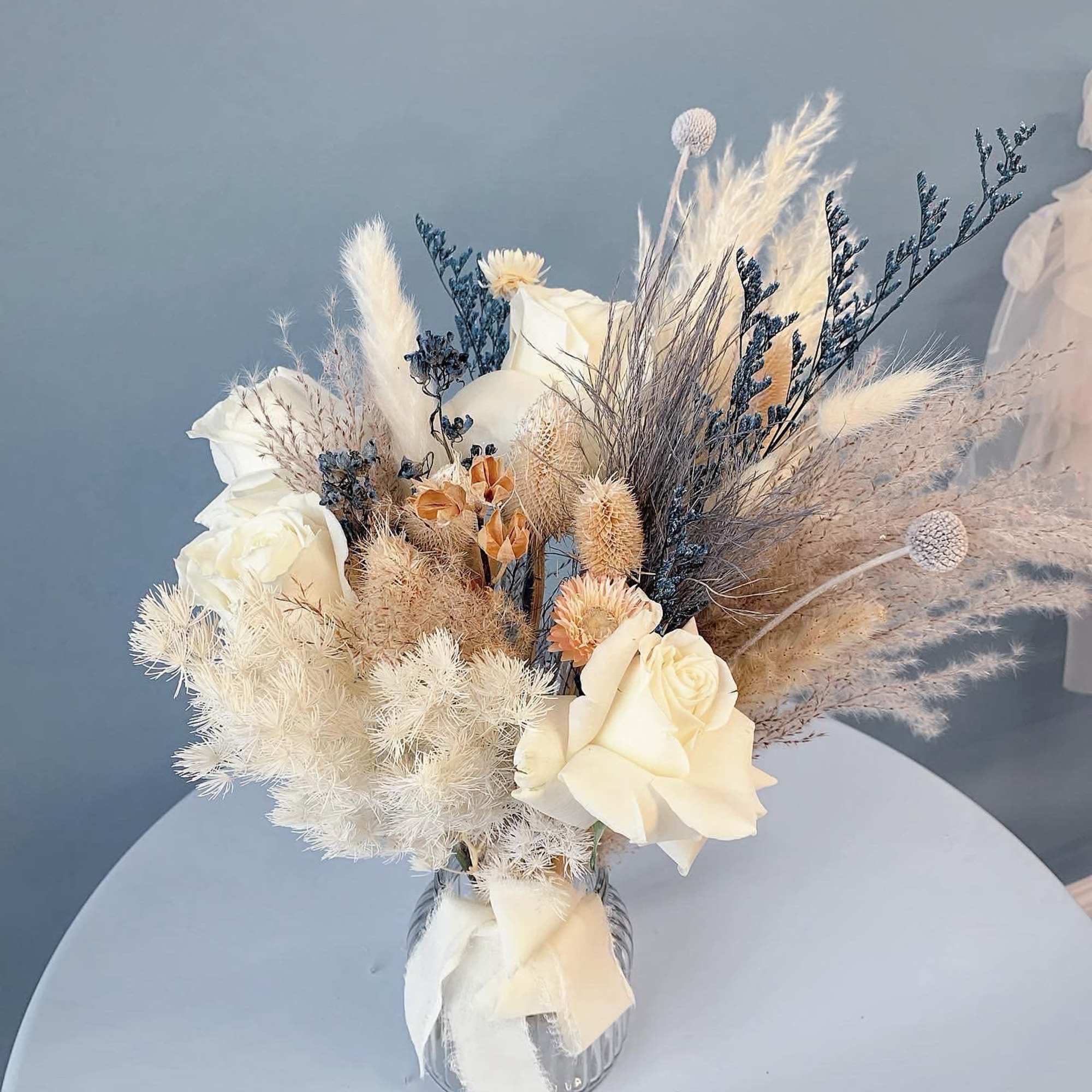 Cream roses and dried grasses arranged in a clear vase with a ribbon