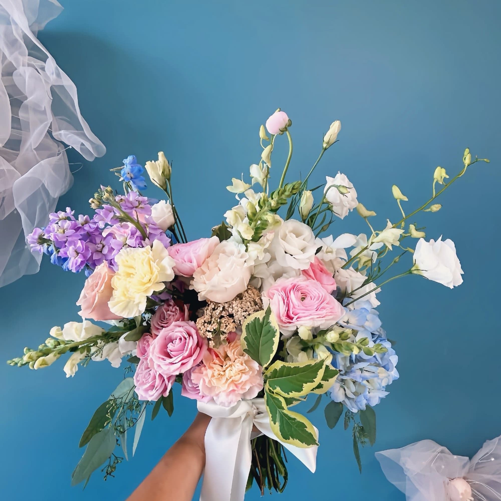 Handheld bouquet of pastel pink, white, blue, and purple flowers tied with a white ribbon