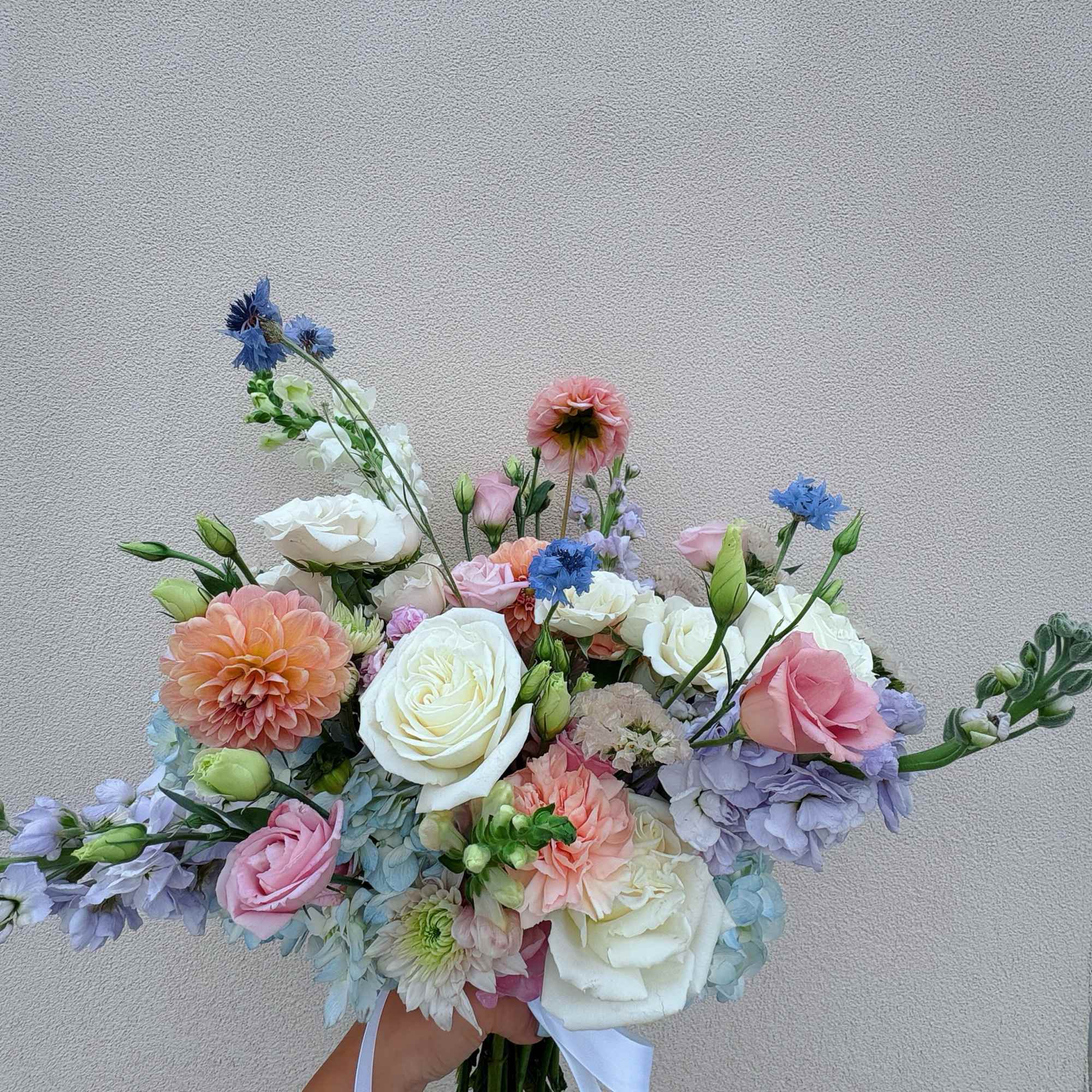 Handheld bouquet of white, pink, peach, and blue flowers with a white ribbon