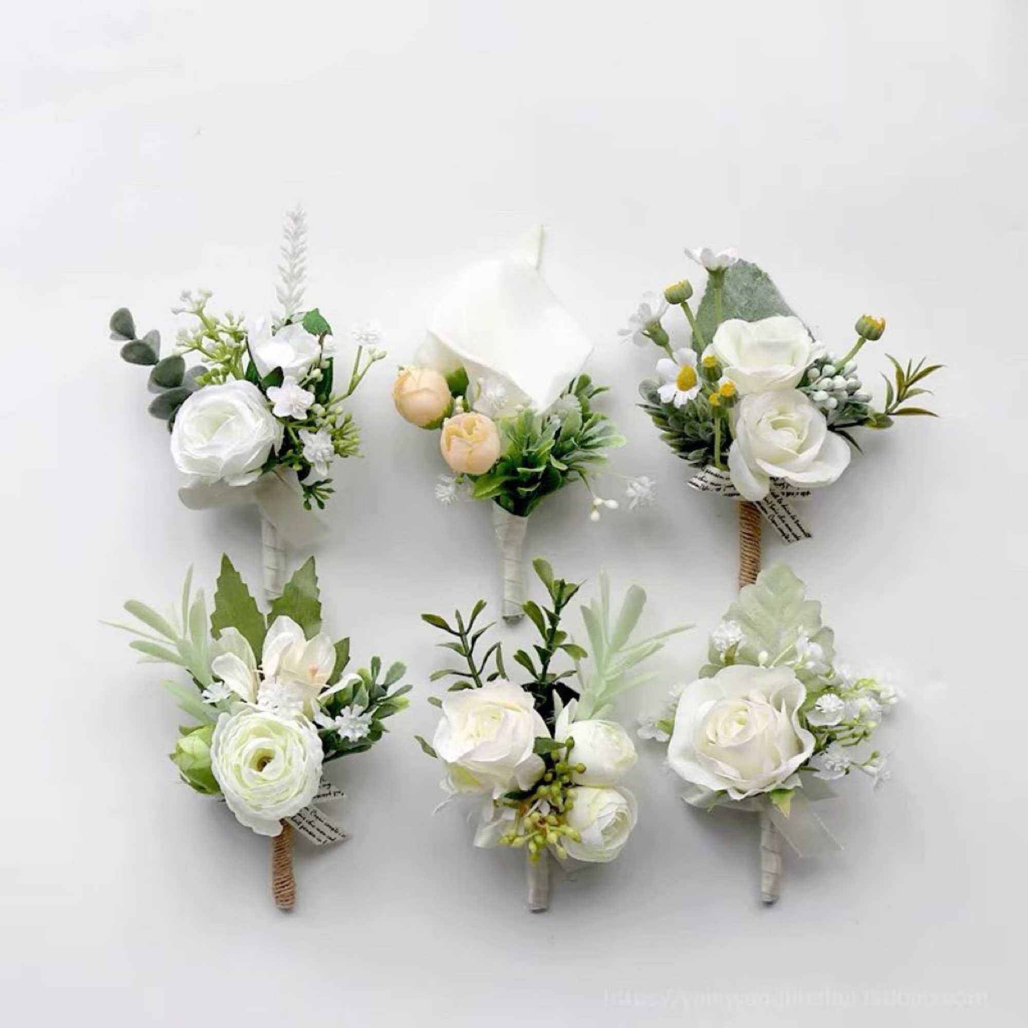 Six small white floral boutonnieres with green accents arranged on a white background