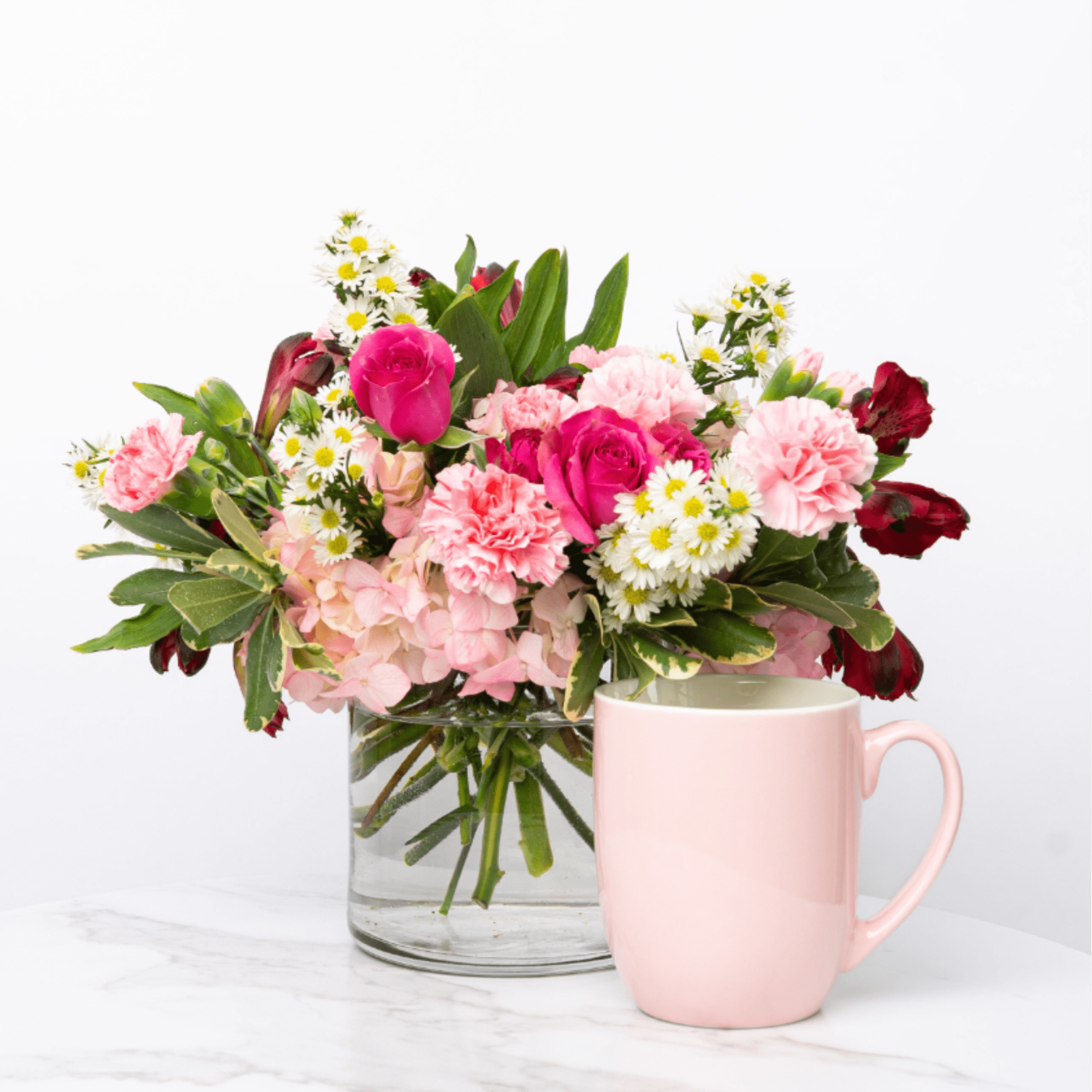 Pink and white mixed flower arrangement in a glass vase beside a pale pink mug