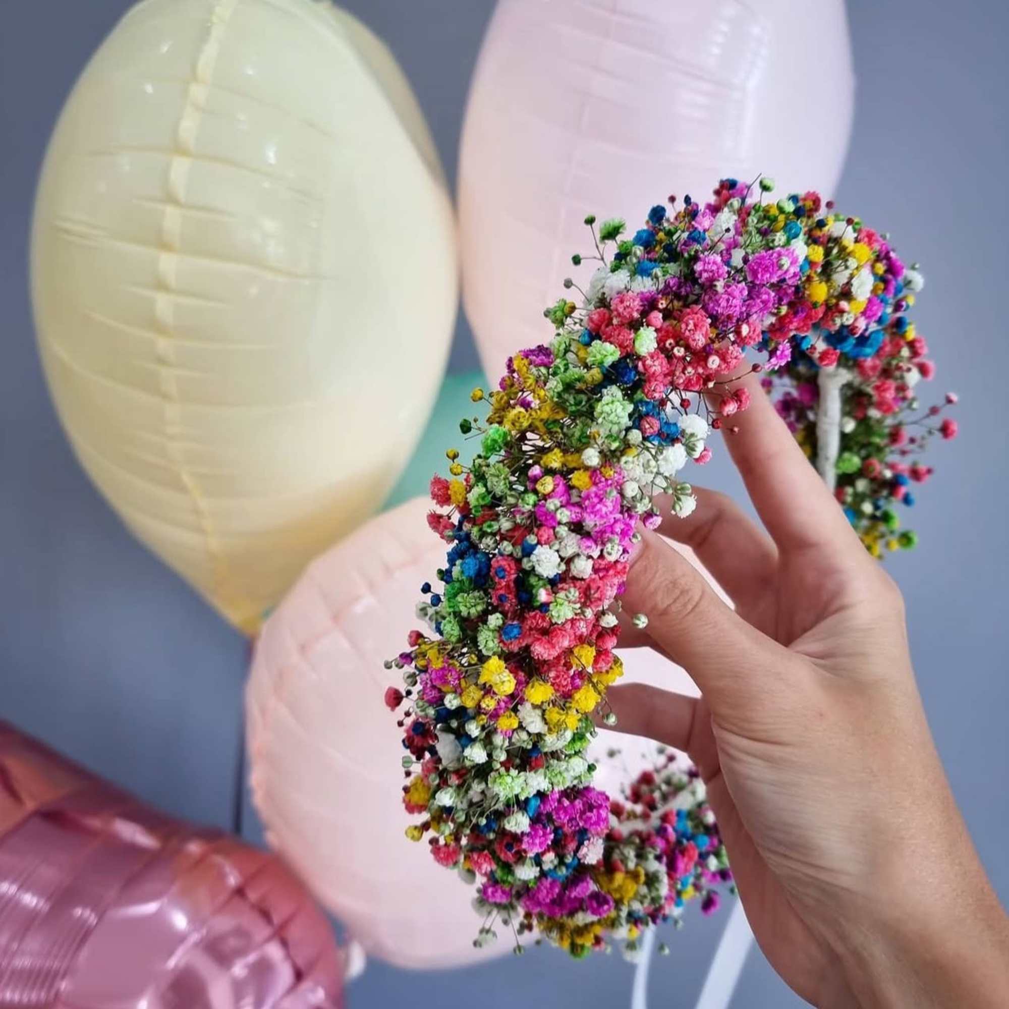 Hand holding a colorful flower crown with pastel balloons in the background