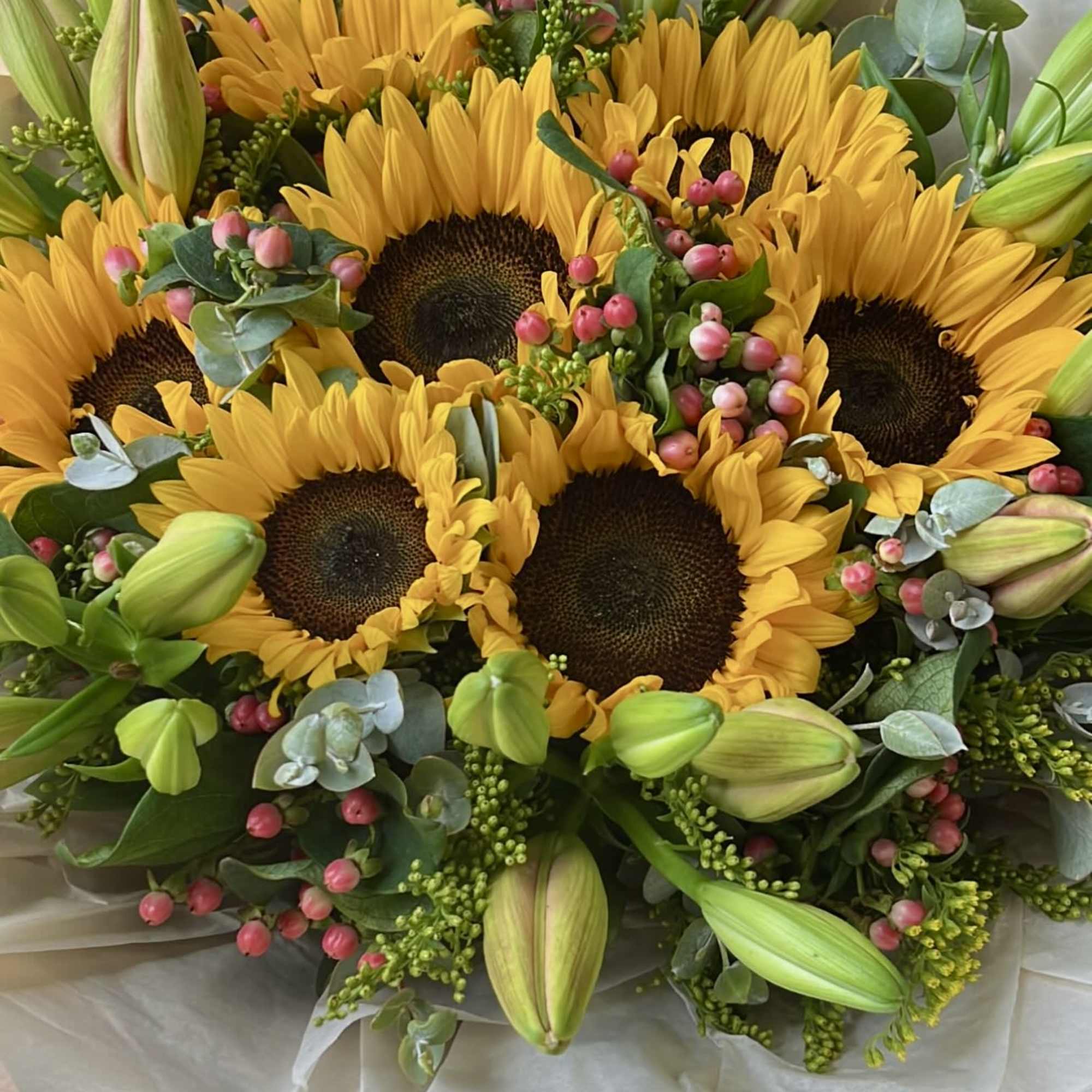 Hand-tied bouquet of yellow sunflowers with green lily buds and pink berries wrapped in cream paper.
