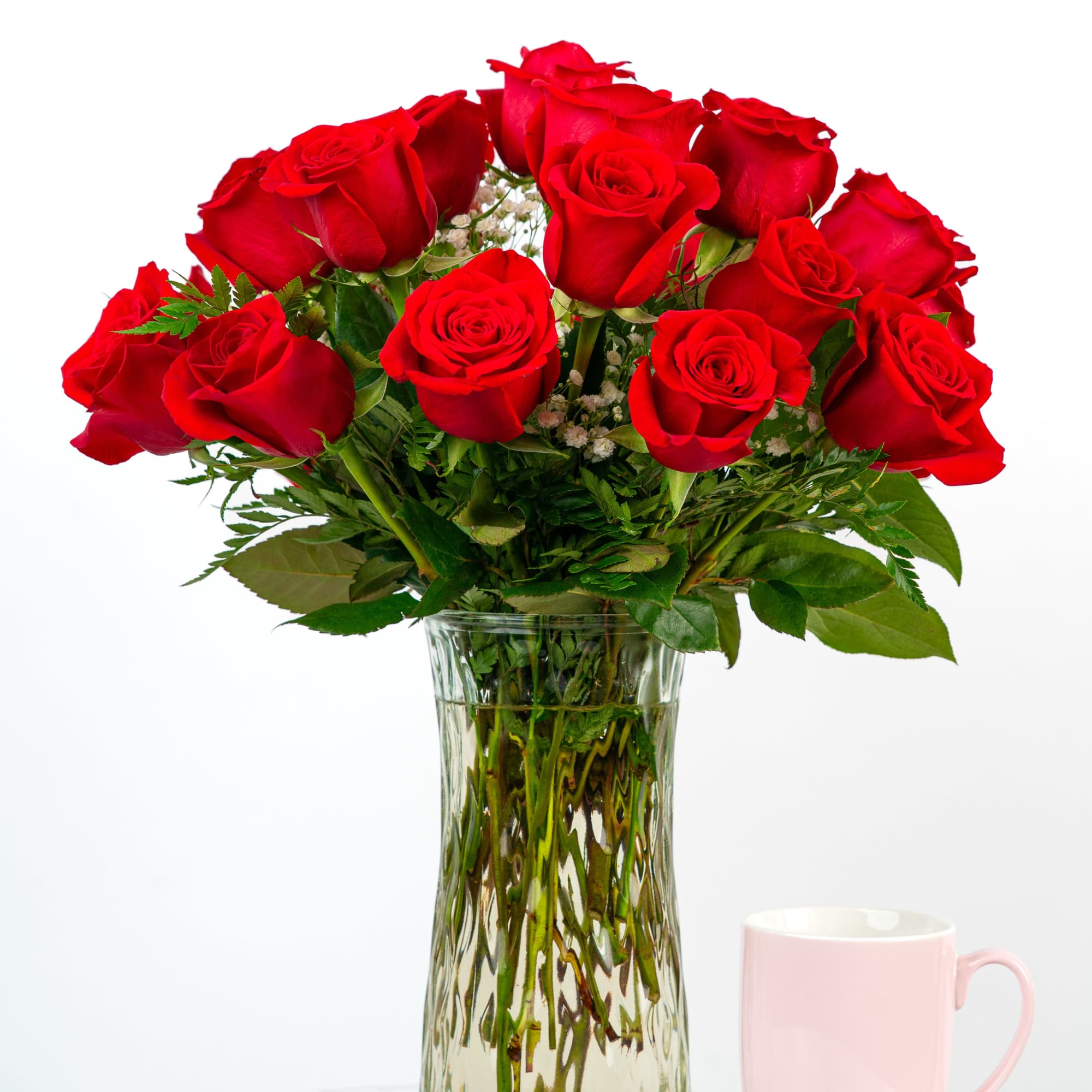 Tall glass vase of red roses with white accents beside a light pink mug