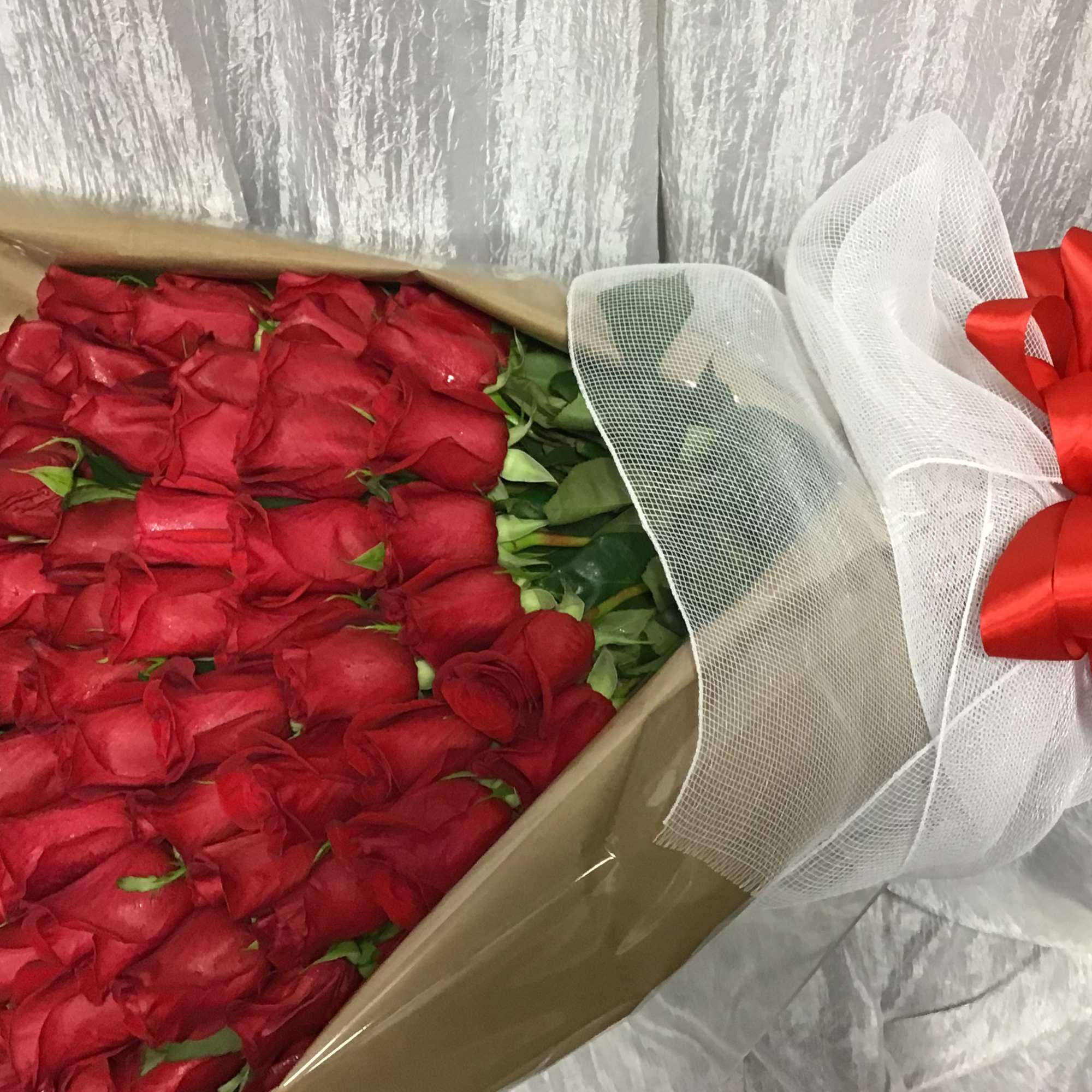 Large bouquet of tightly packed red roses wrapped in kraft paper with white mesh and a red bow.
