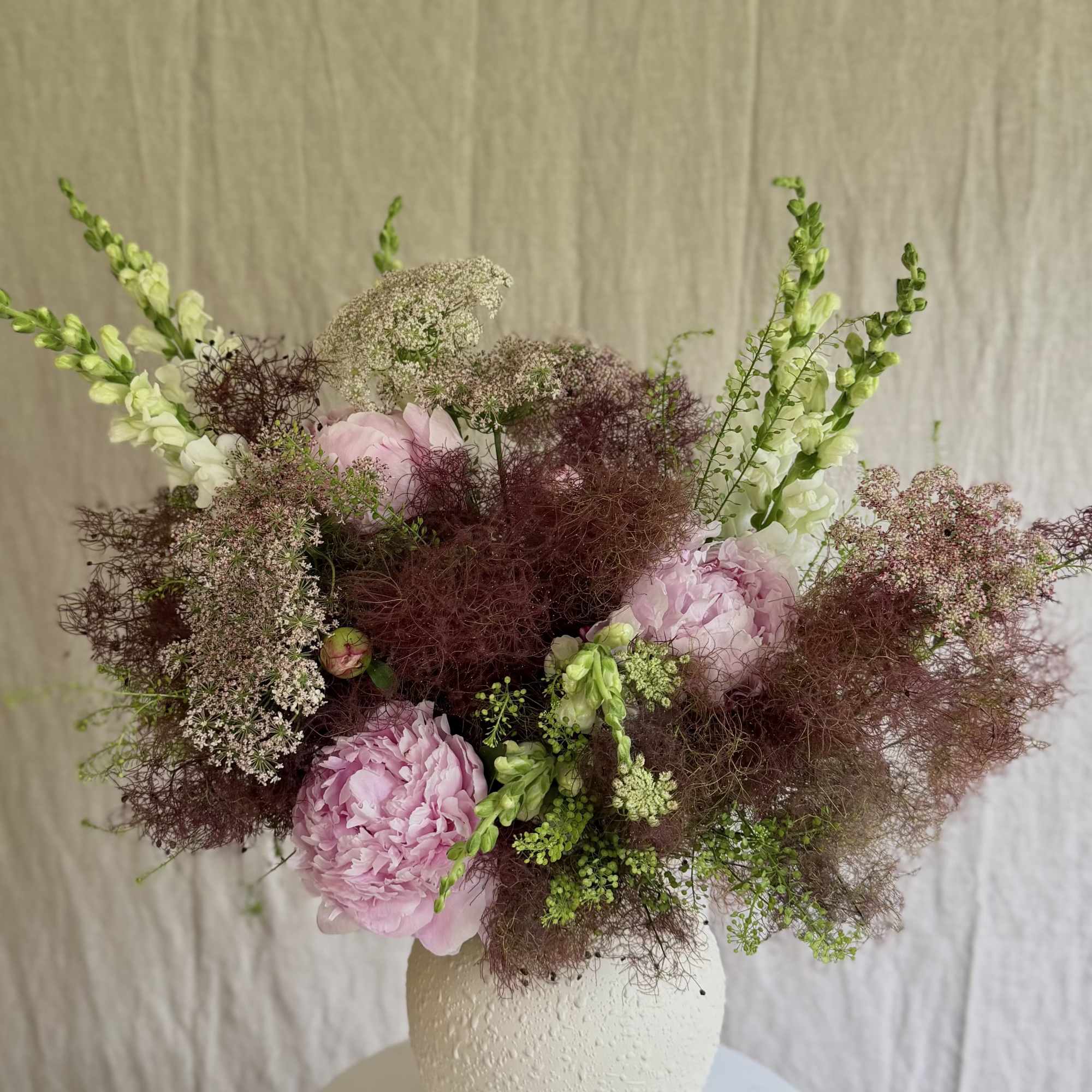 Soft blush peonies and tall white snapdragon spikes mingle with airy burgundy