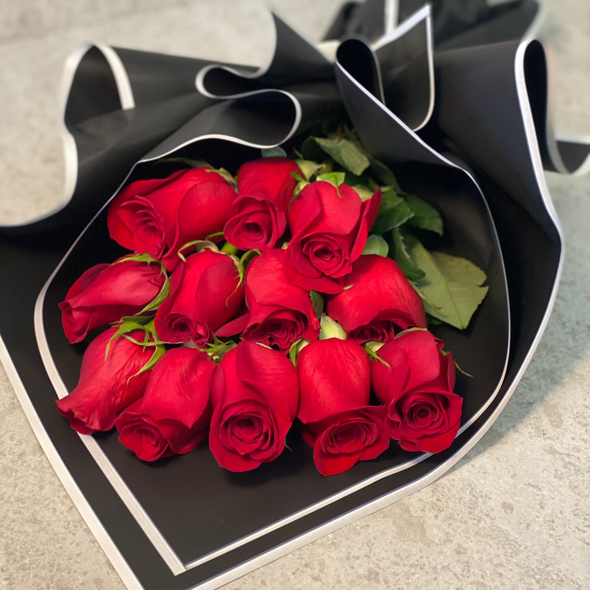 Dozen red roses wrapped in black paper with white trim on a stone surface