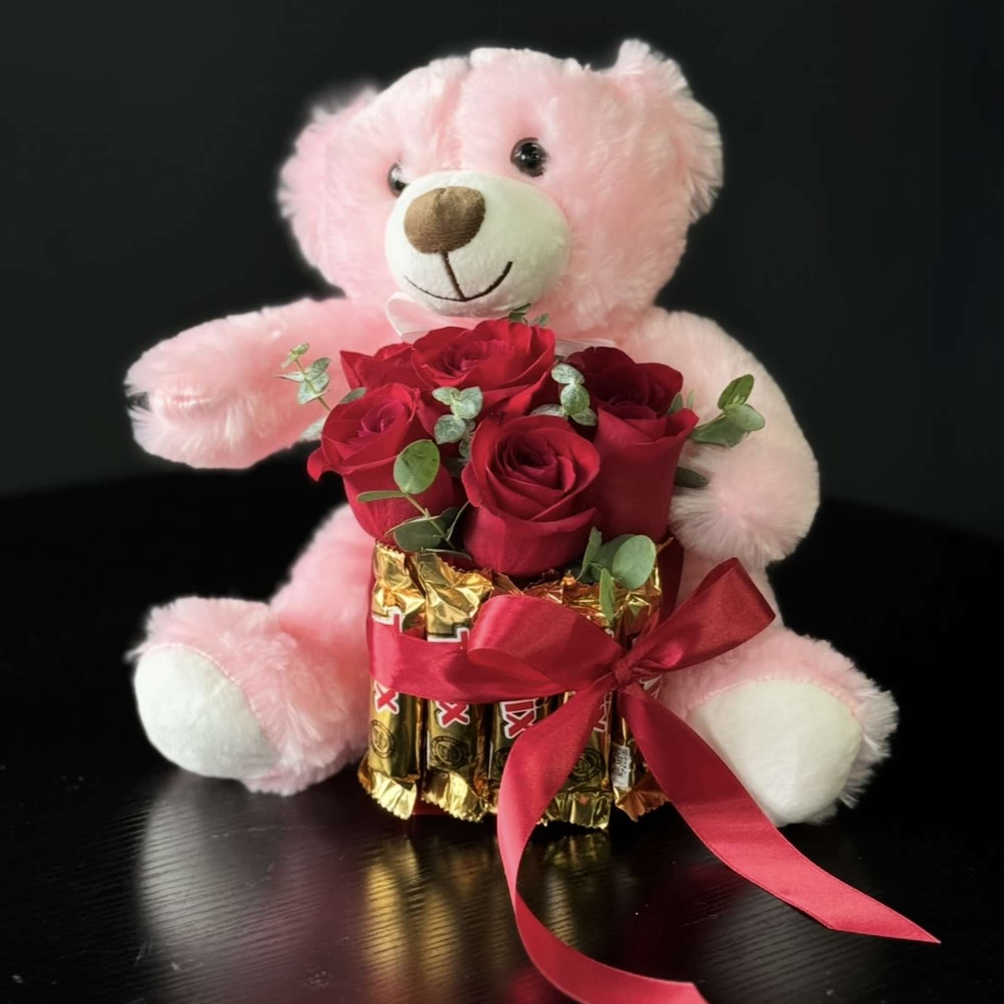 Pink teddy bear holding red roses and chocolates with a ribbon