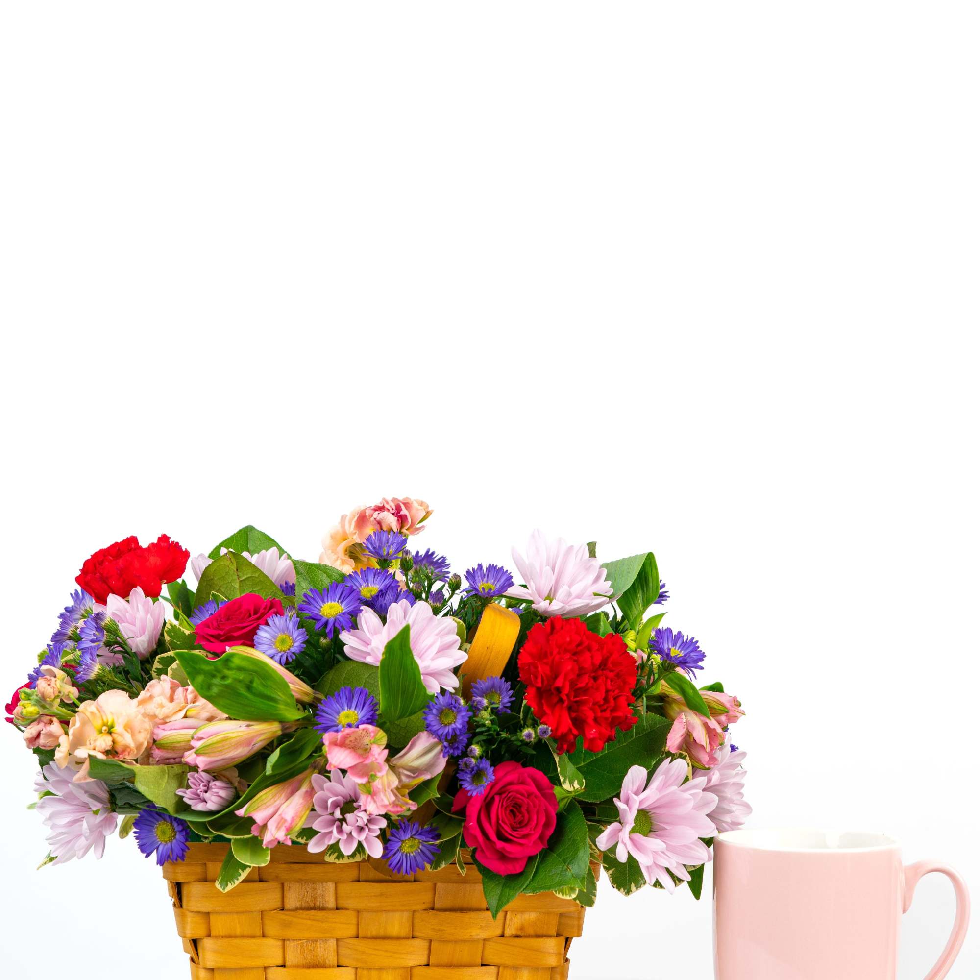 Colorful mixed flower basket arrangement beside a pink mug on a white surface.