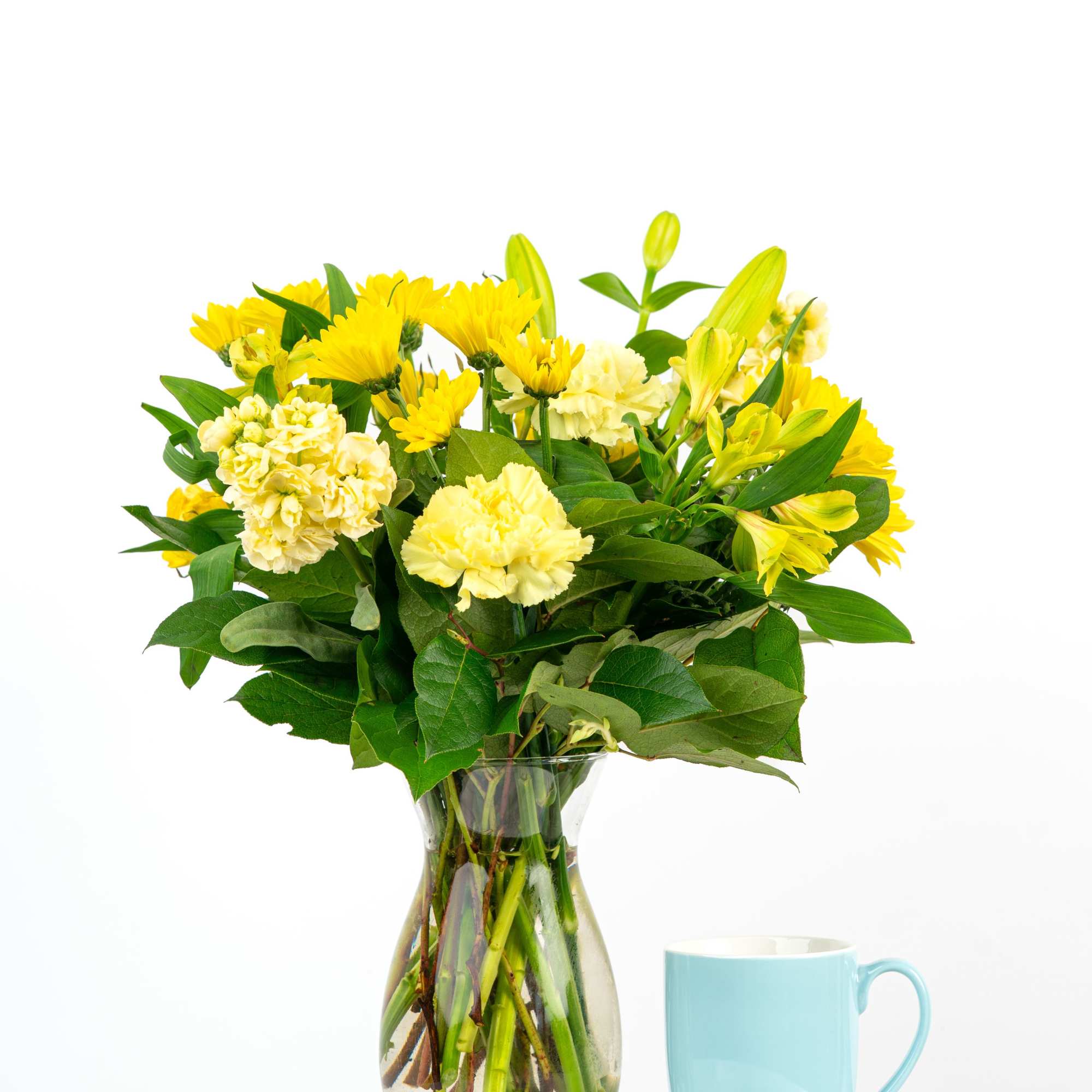 Yellow and cream flowers in a clear glass vase beside a light blue mug on a white surface