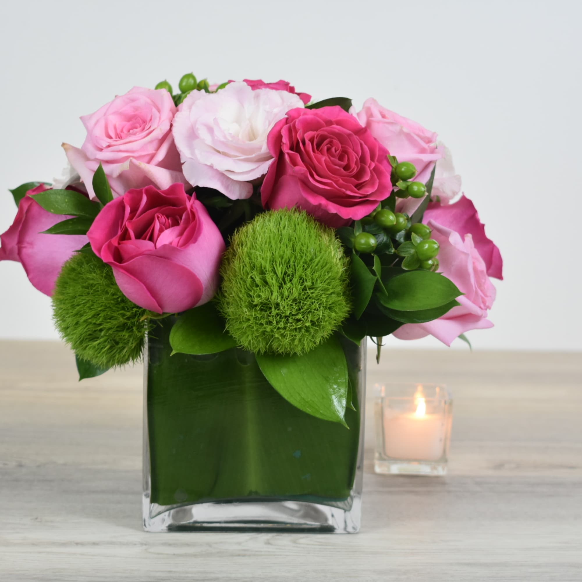 Short arrangement of pink roses and green dianthus balls in a square glass vase with a small candle beside it