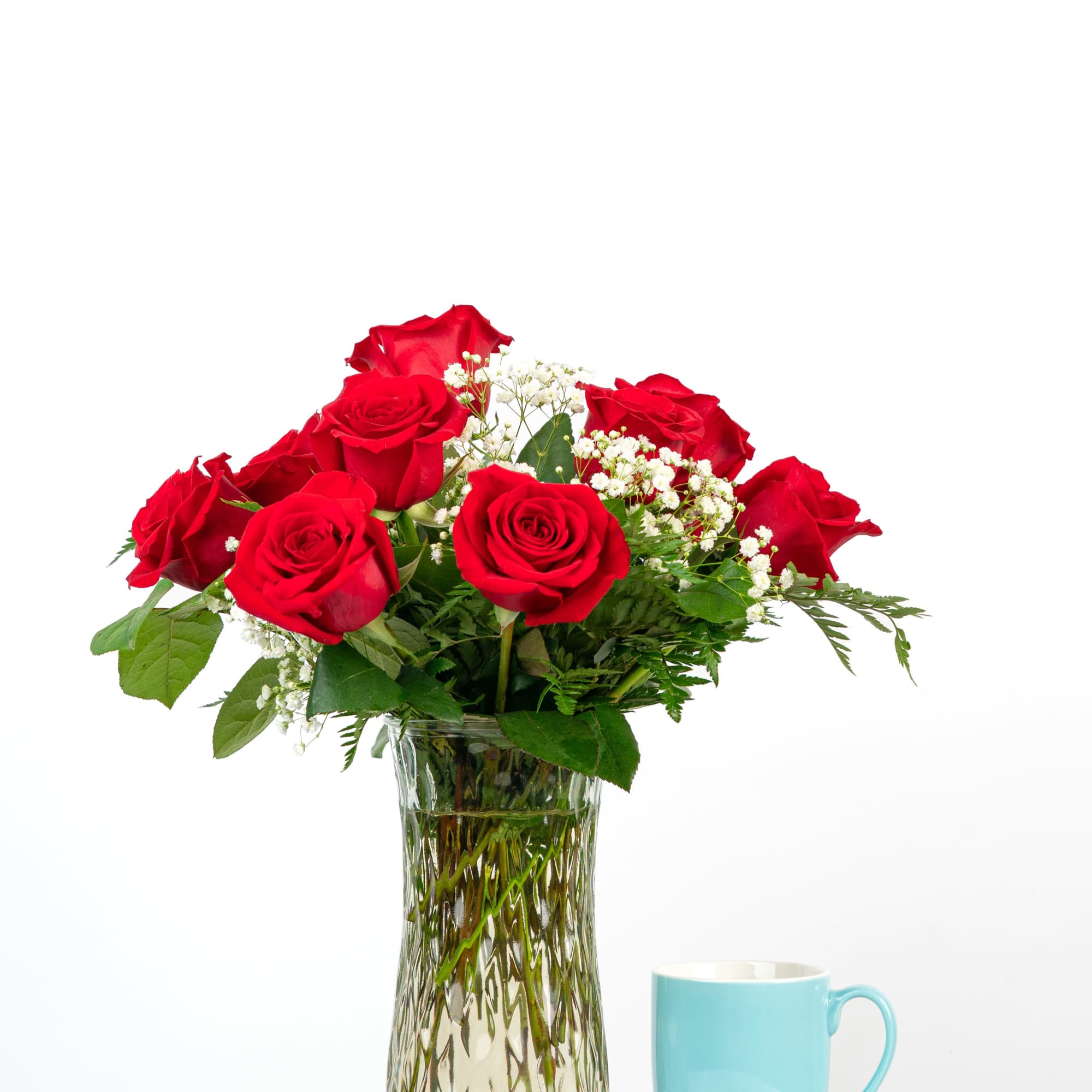 Red roses arranged in a clear glass vase beside a light blue mug