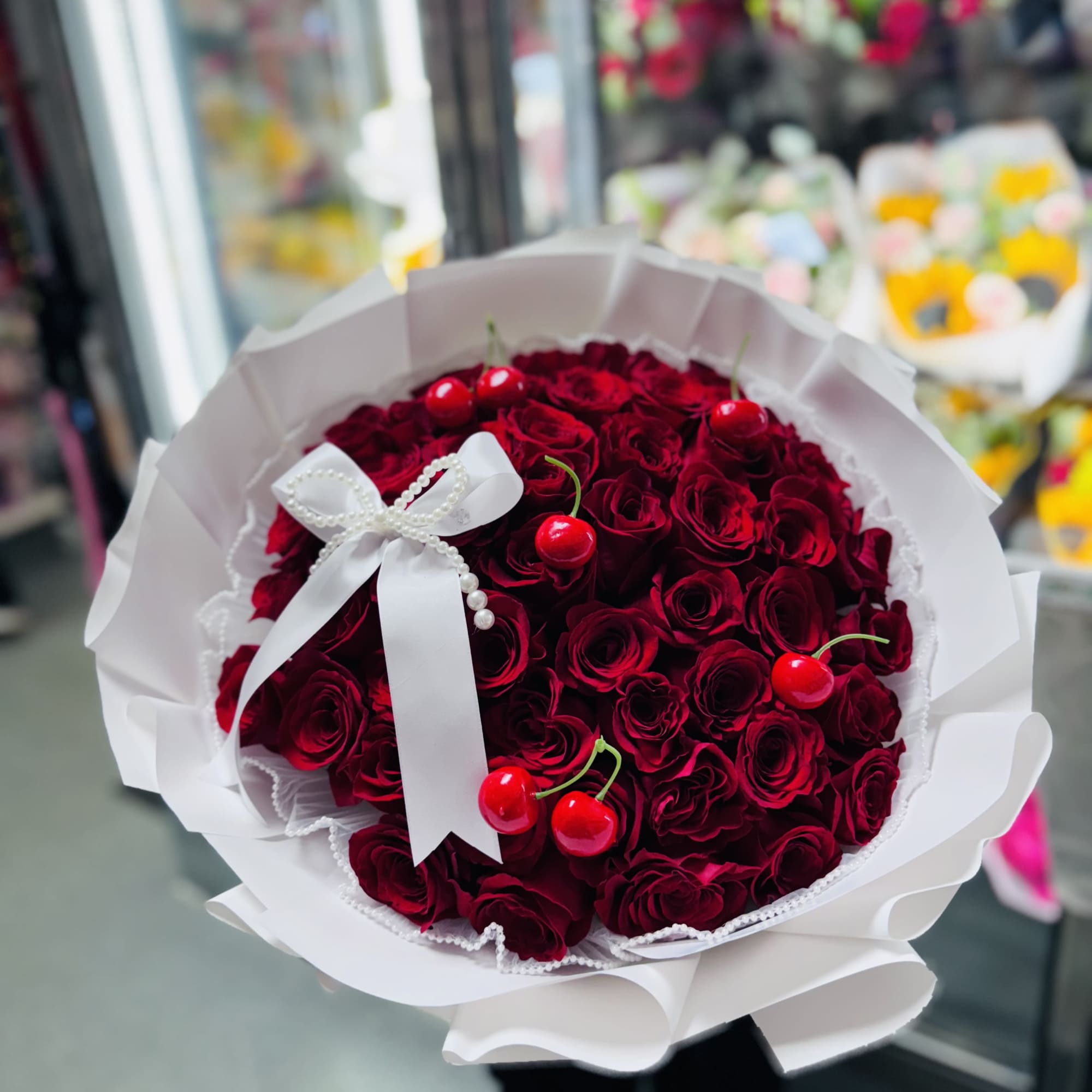 Round bouquet of deep red roses with decorative cherries, wrapped in white paper with a white bow
