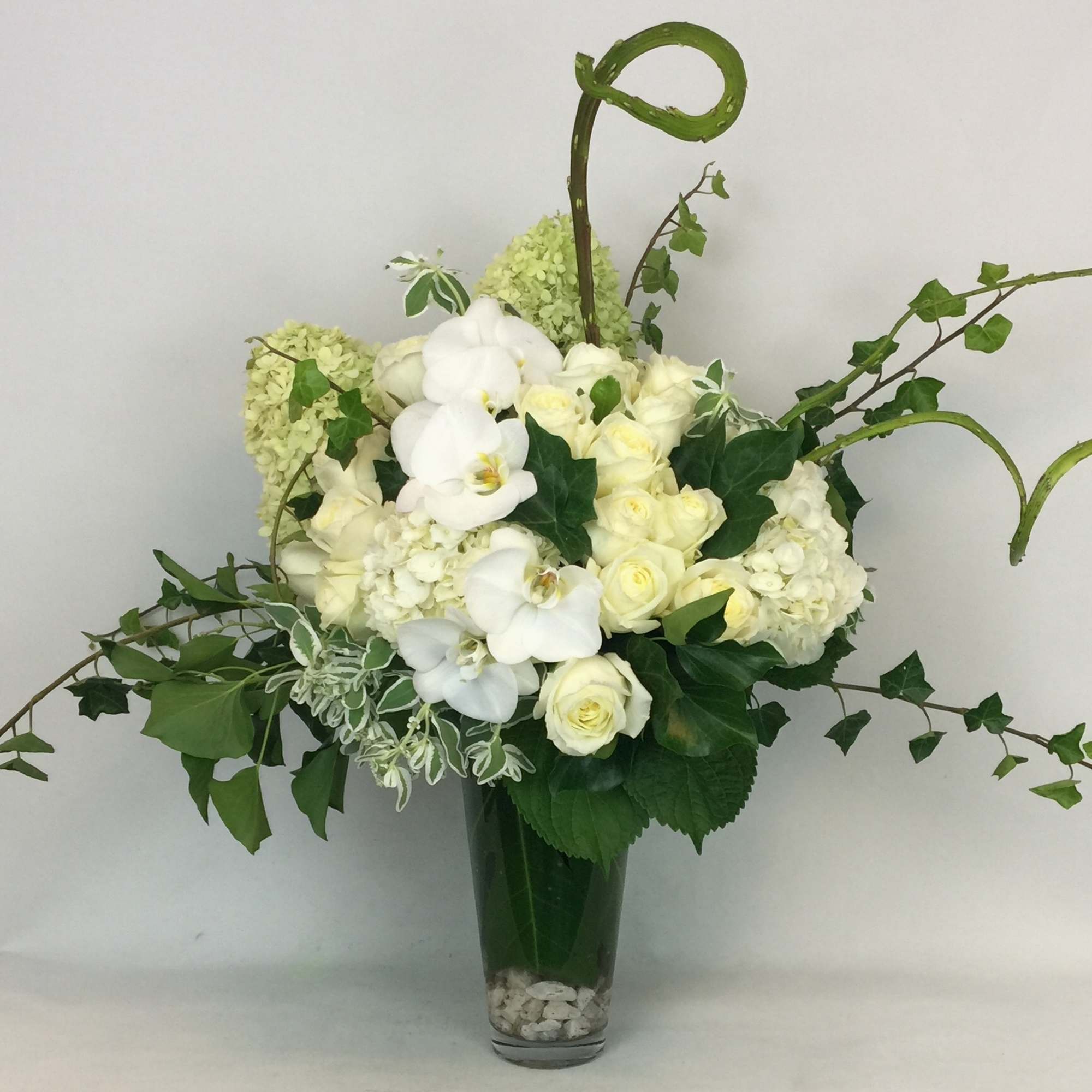 White orchids and roses in a tall glass vase with trailing greenery