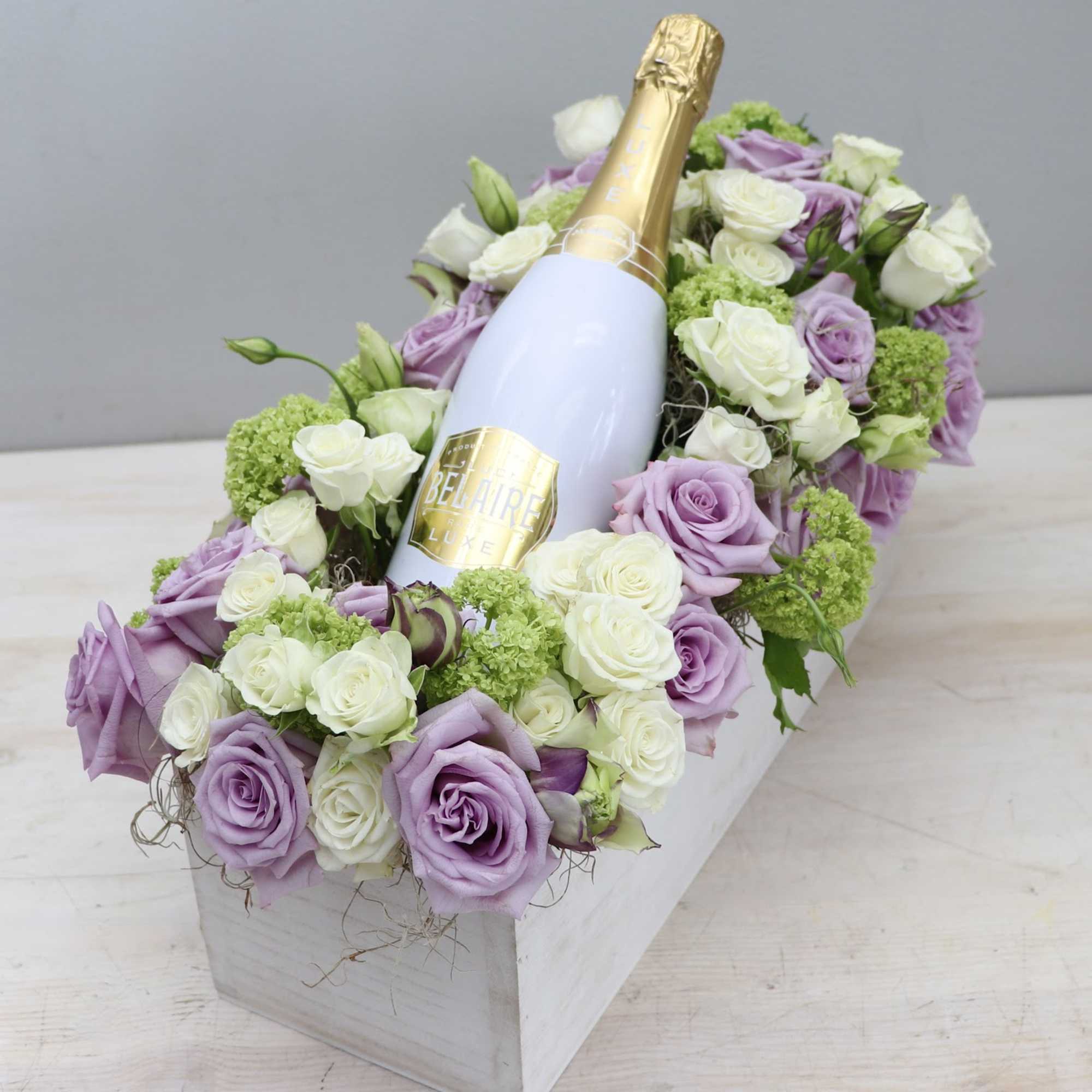 Rectangular box arrangement of lavender and white roses surrounding a champagne bottle