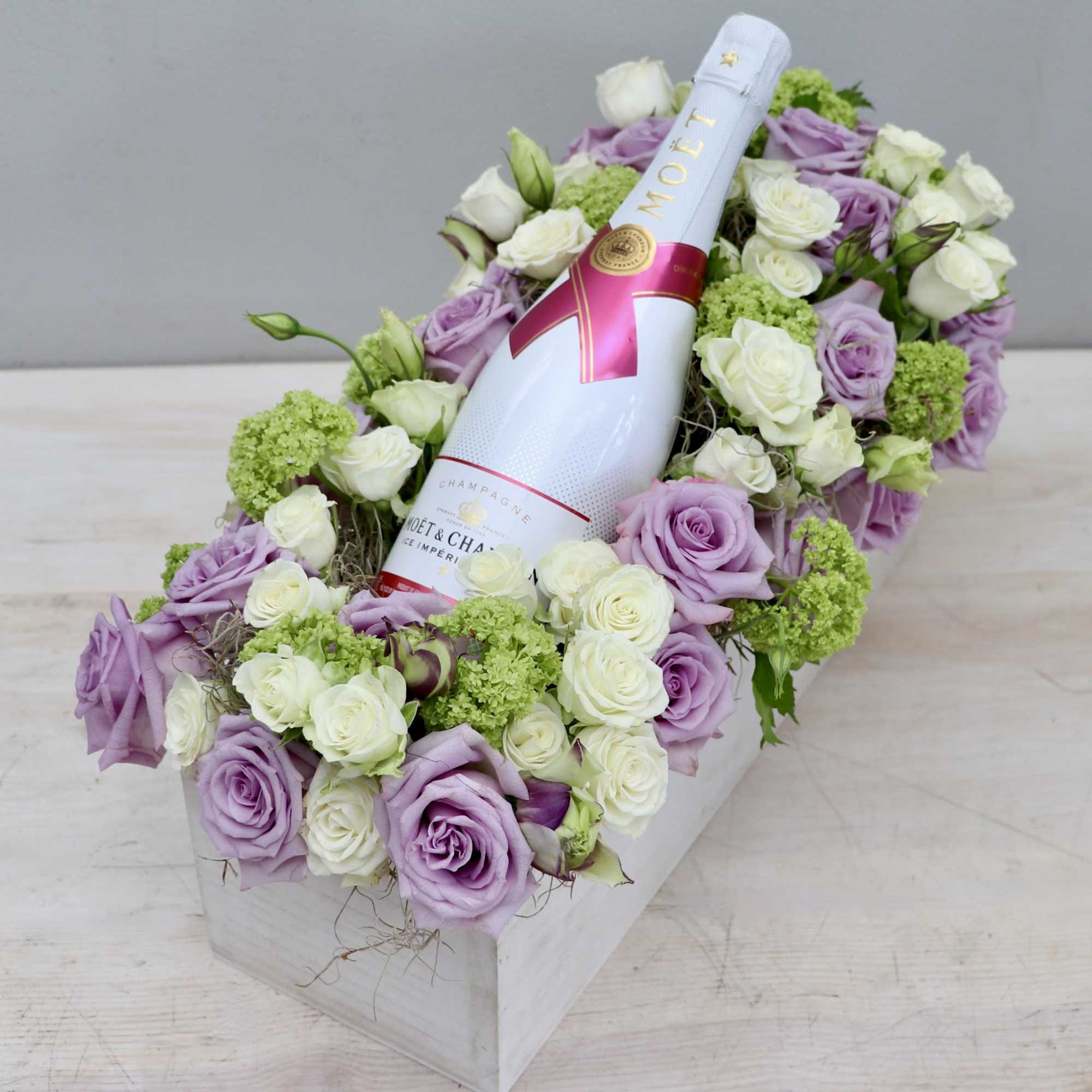 Rectangular box arrangement of lavender and white roses with green blooms cradling a white champagne bottle.