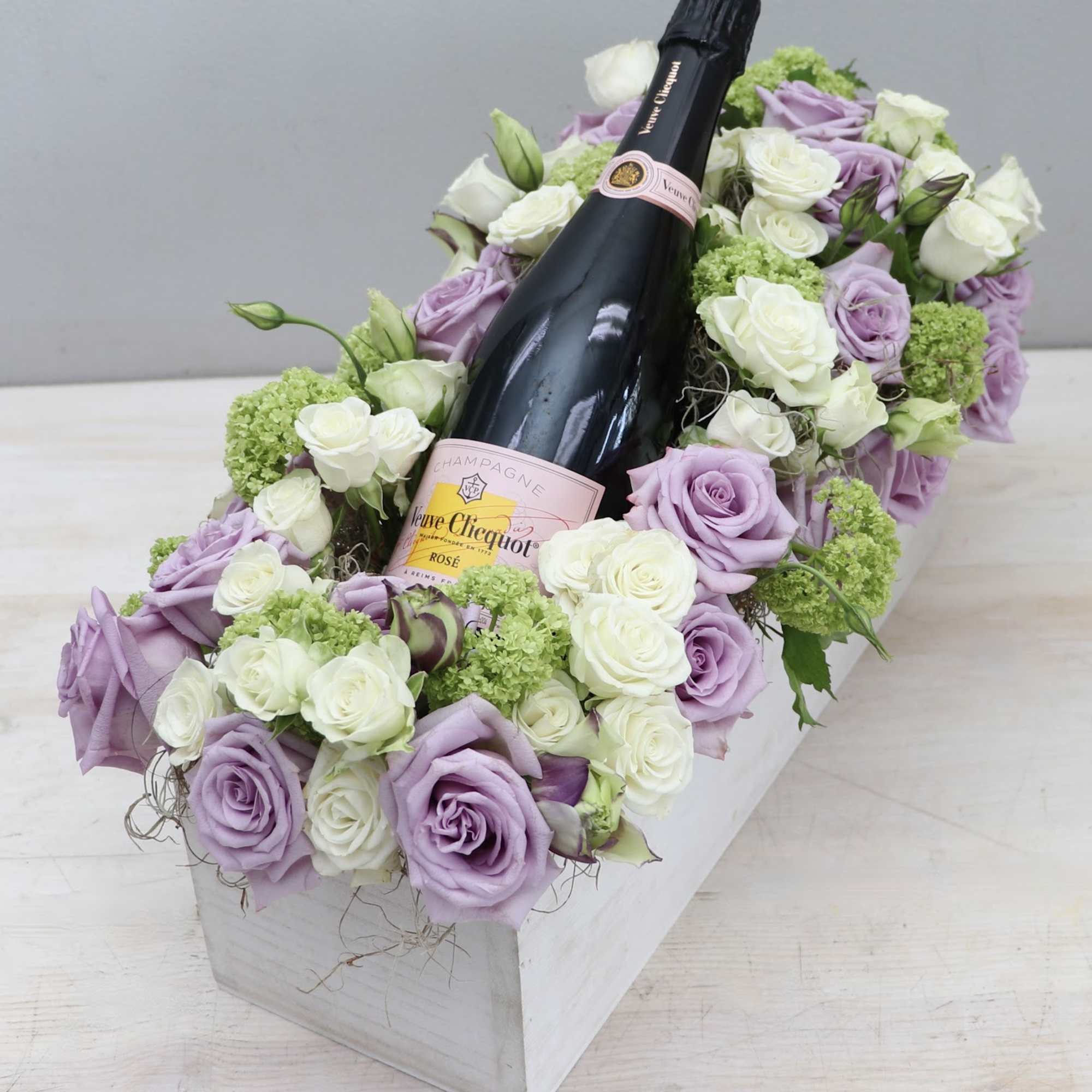 Rectangular box of lavender and white roses surrounding a rosé champagne bottle