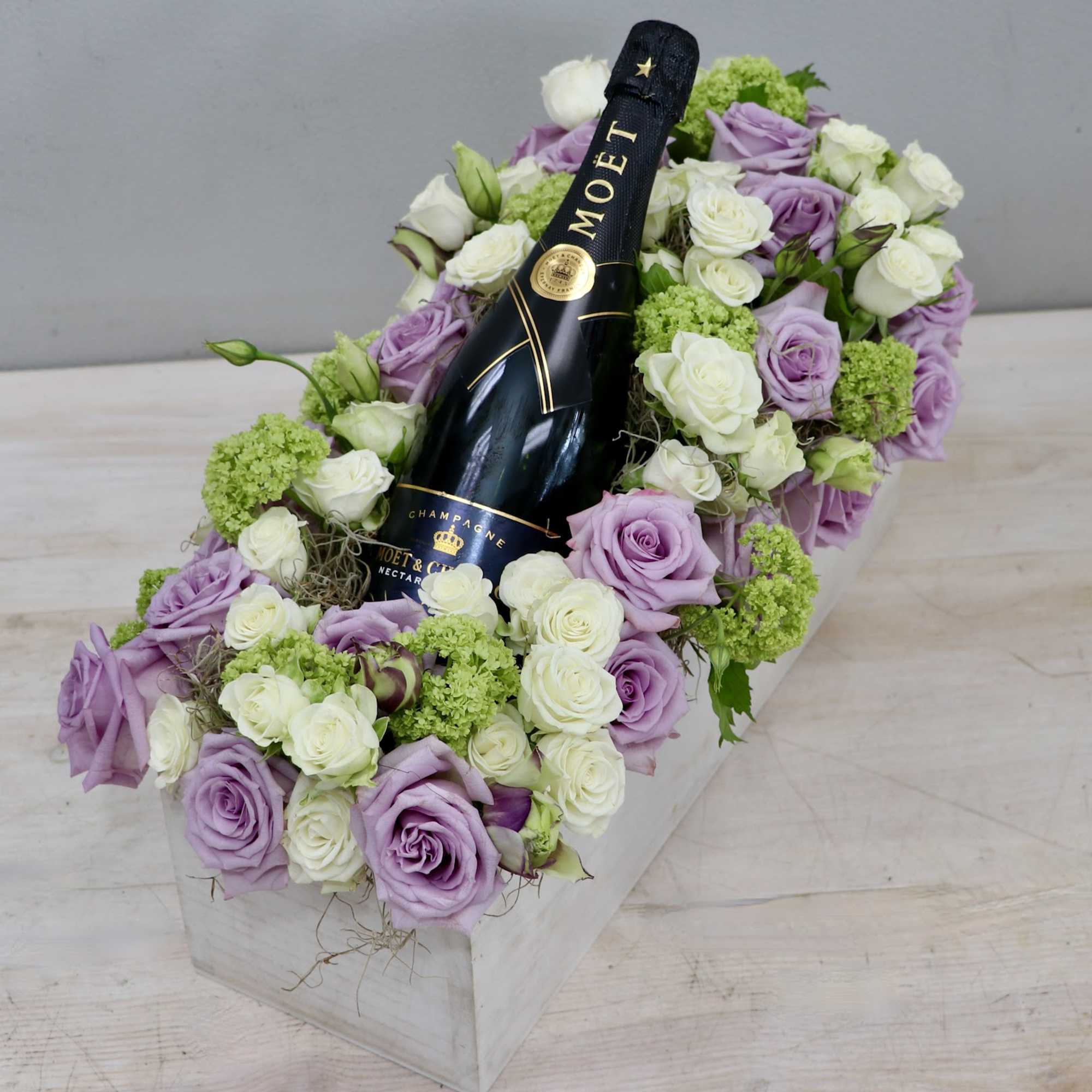 Rectangular box of lavender and white roses cradling a bottle of champagne.