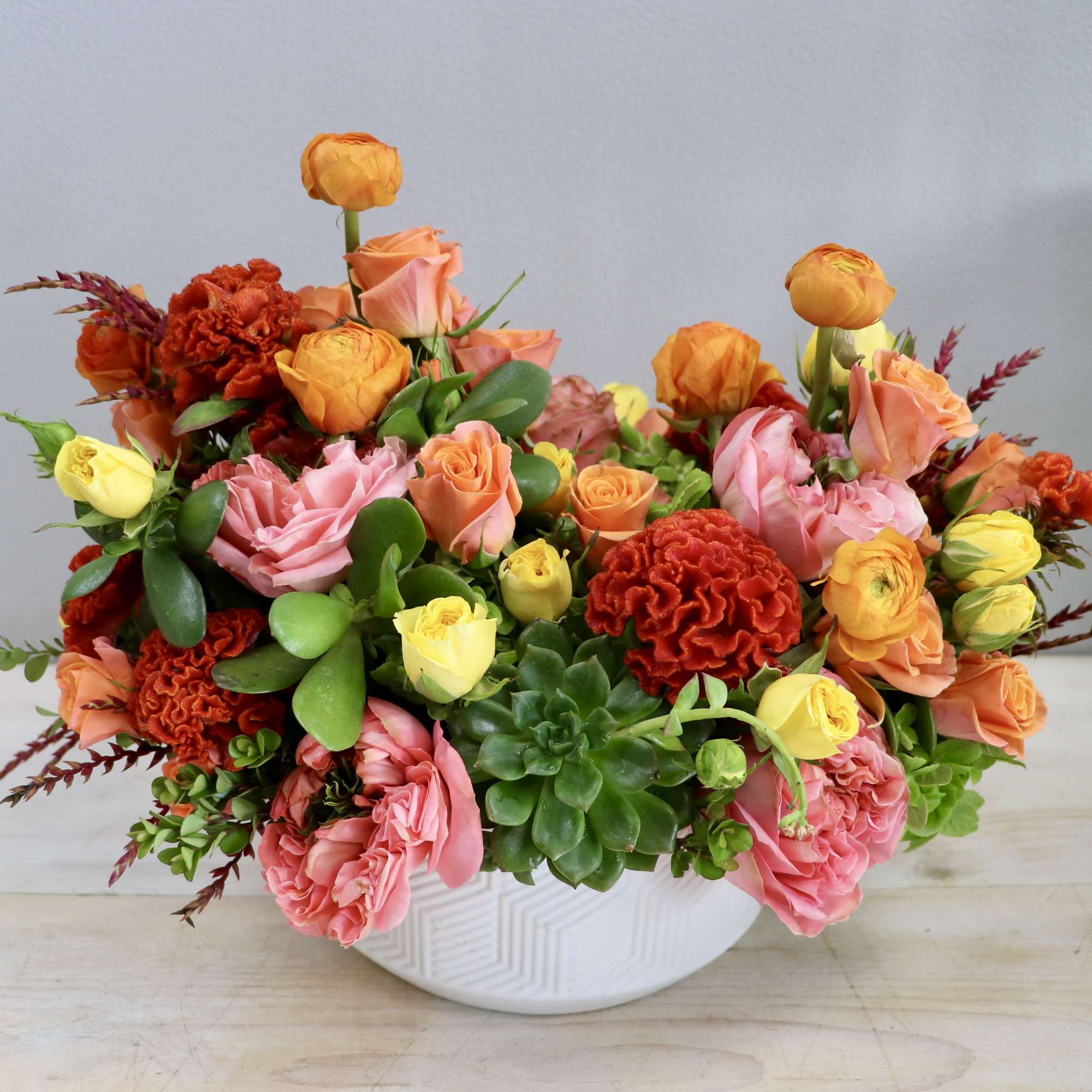 Low arrangement of orange, pink, and yellow roses, ranunculus, carnations, and succulents in a white ceramic bowl