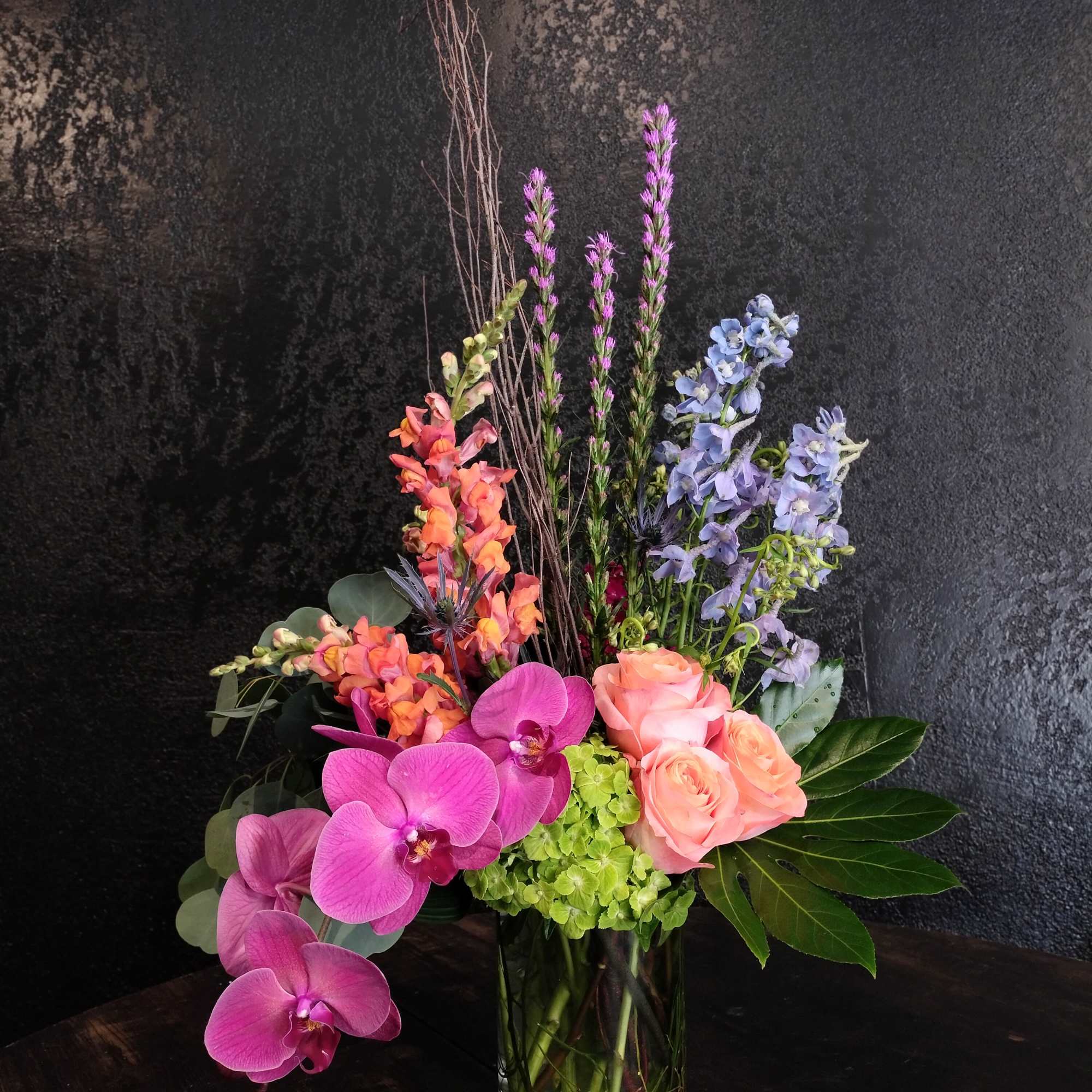 Tall modern arrangement with pink orchids, peach roses, blue delphinium and green hydrangea in a glass vase