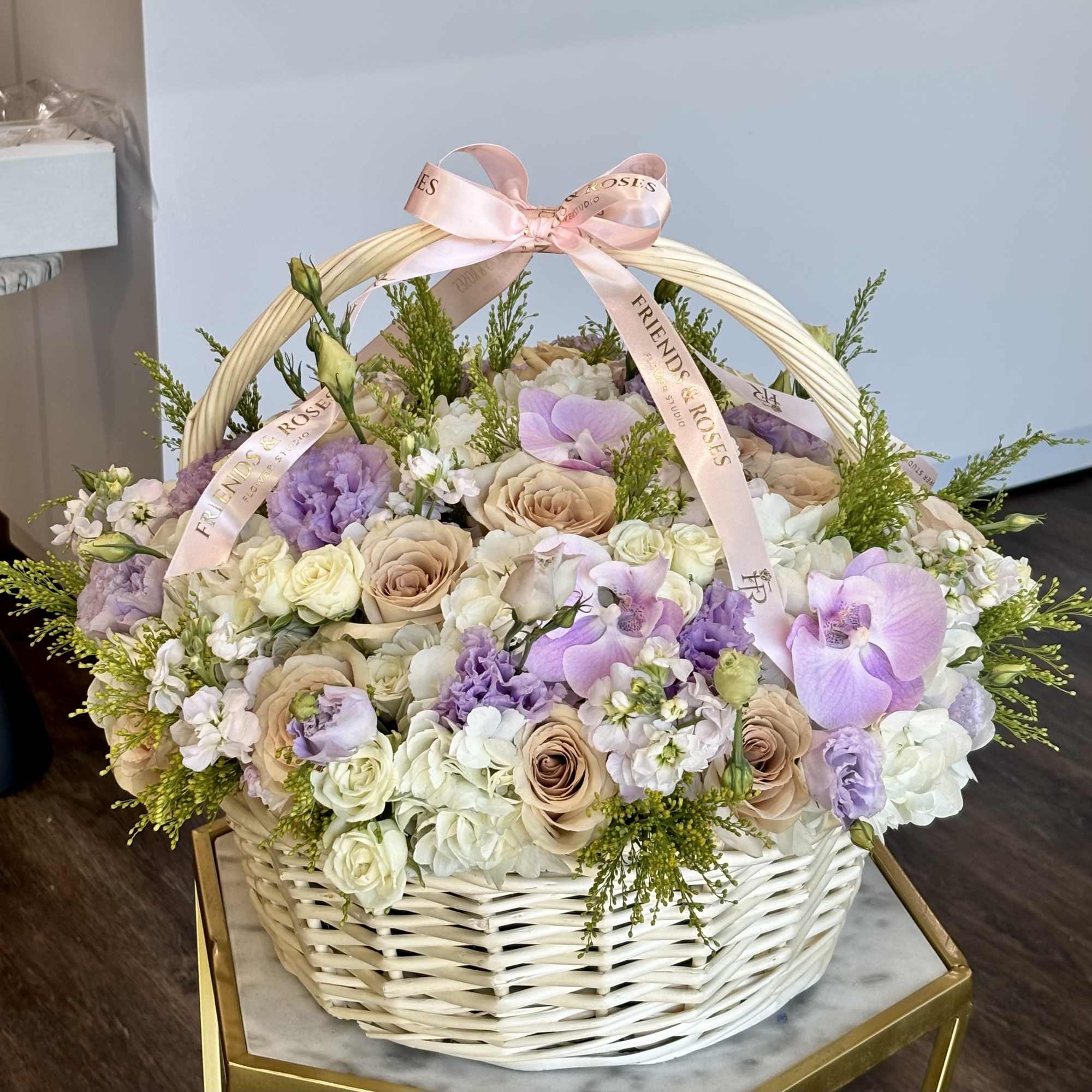 Basket arrangement of pastel roses, orchids, and white flowers with a pink ribbon handle