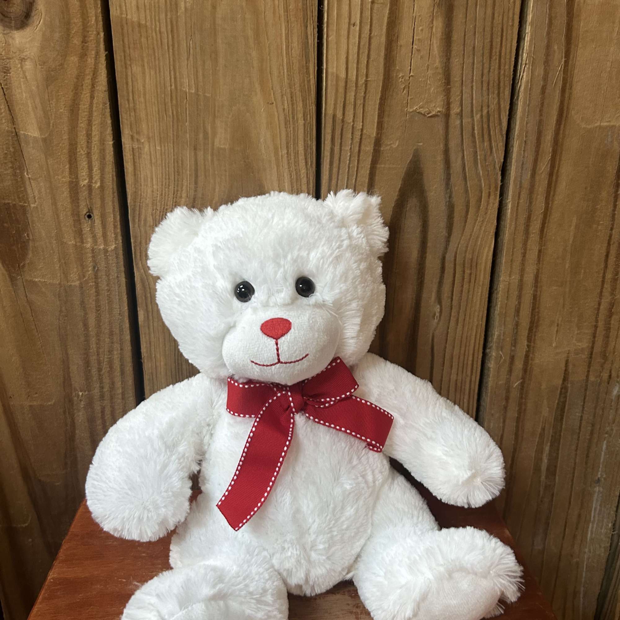 White plush teddy bear with a red bow sitting on a wooden surface against wood planks.