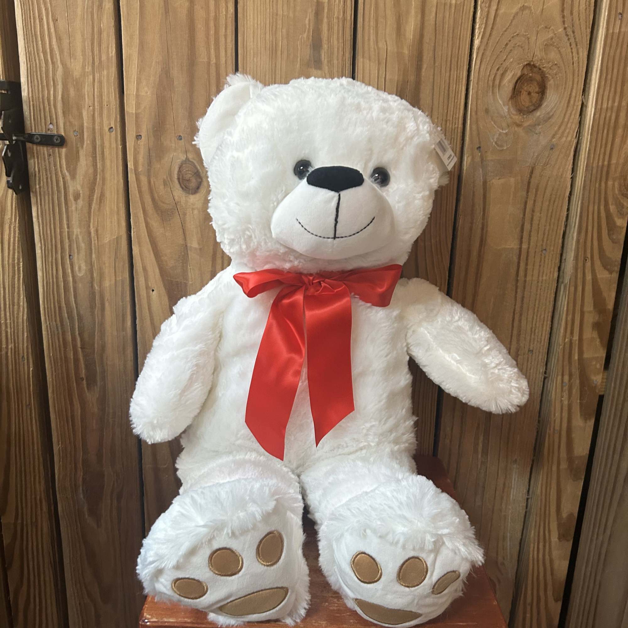 White plush teddy bear with a red satin bow sitting on a wooden stool.