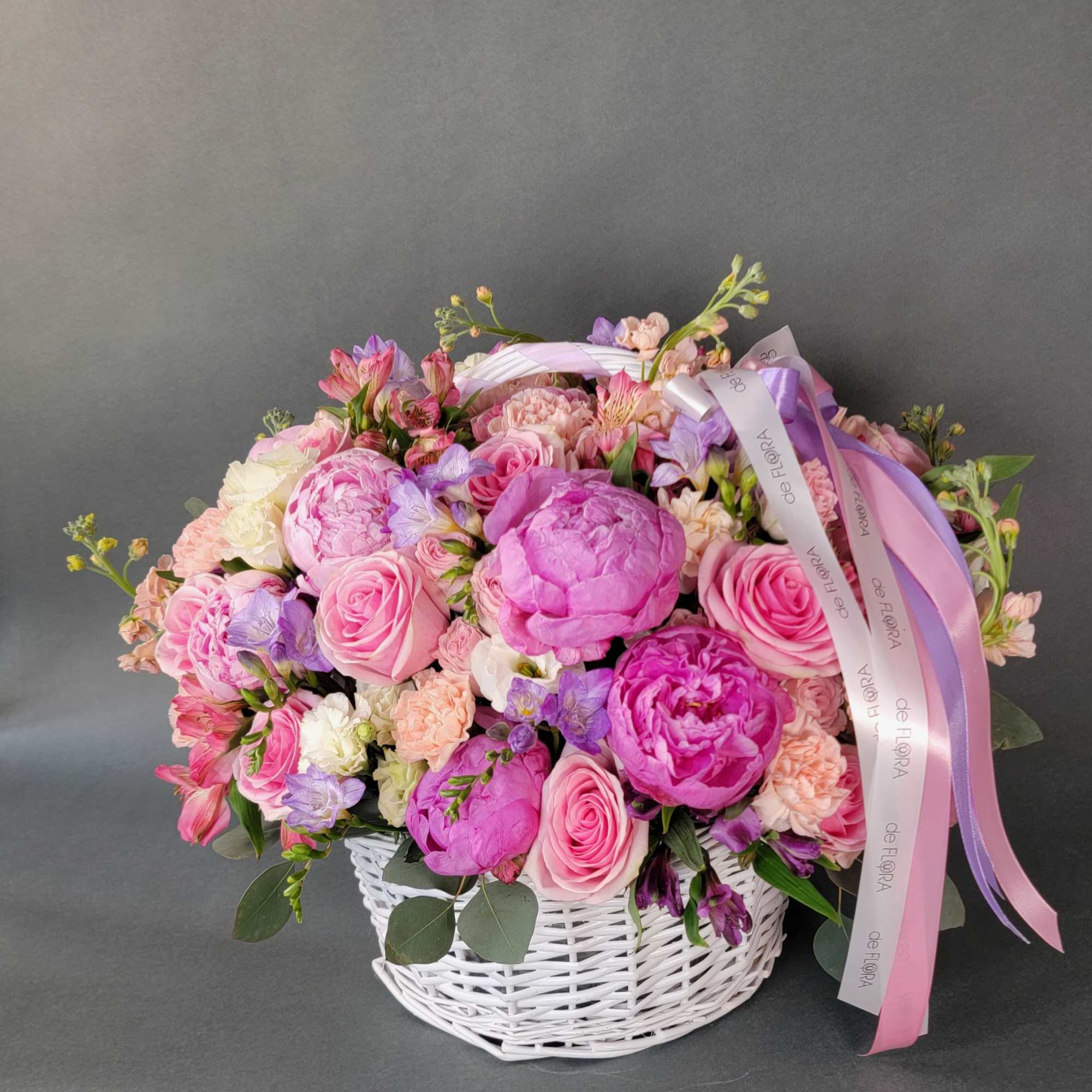 Pink and lavender flower arrangement in a white wicker basket with ribbon