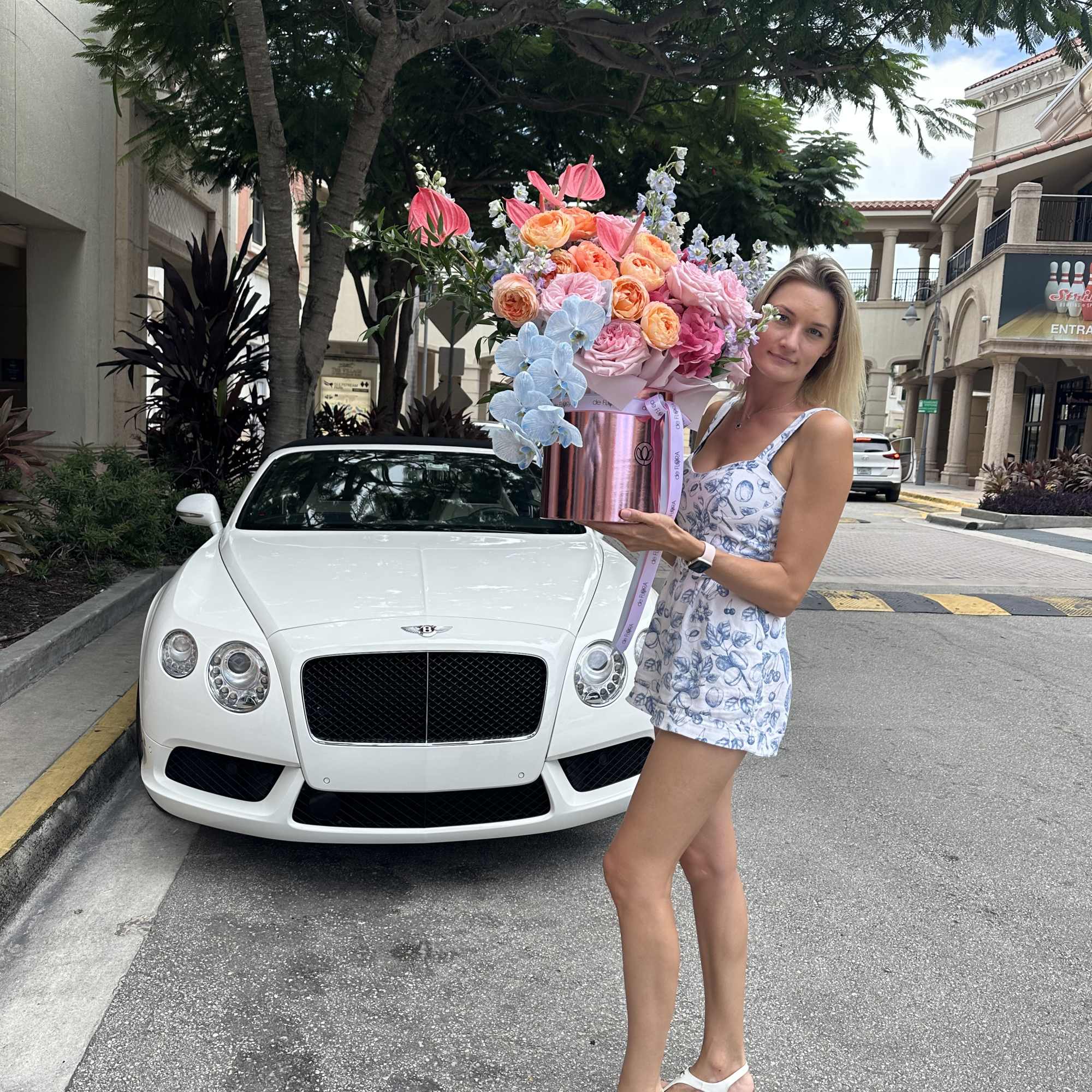 Woman holding a large pastel flower arrangement in a pink box beside a white car