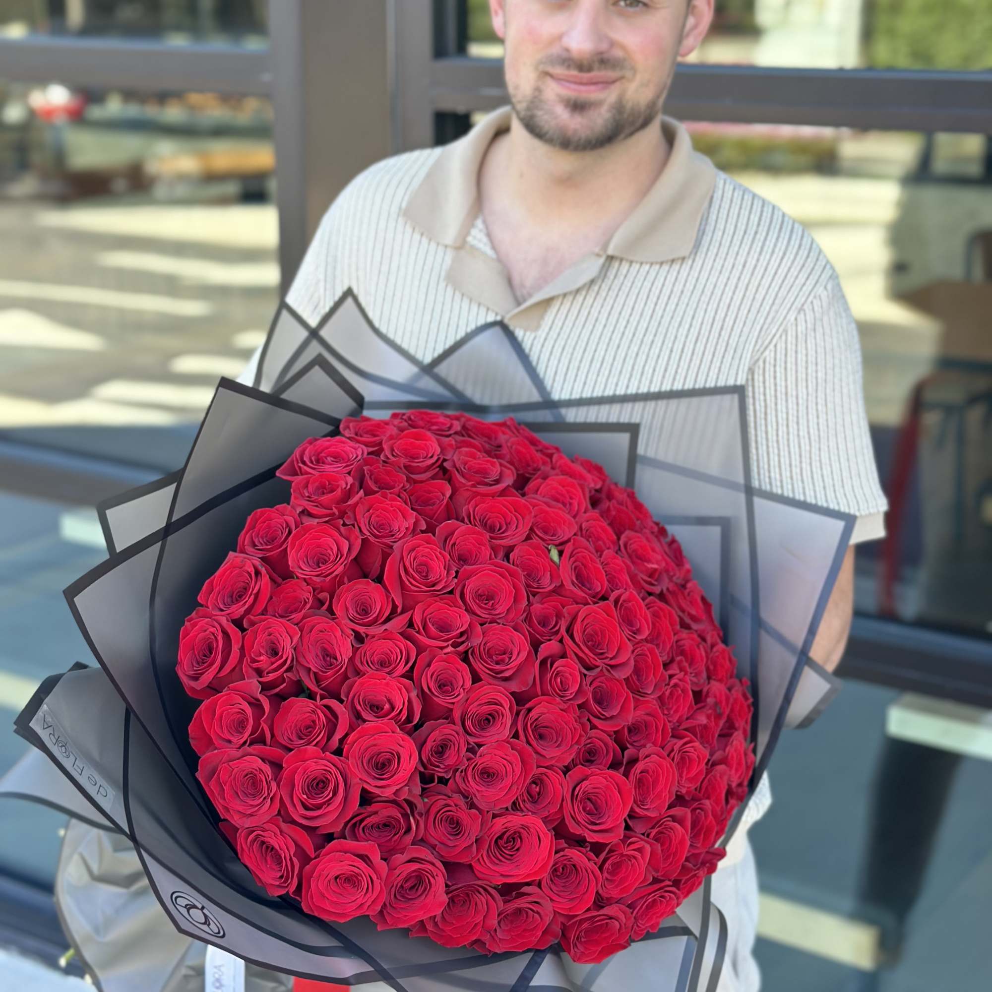 Large round bouquet of tightly packed red roses wrapped in black-edged translucent paper, held by a person.
