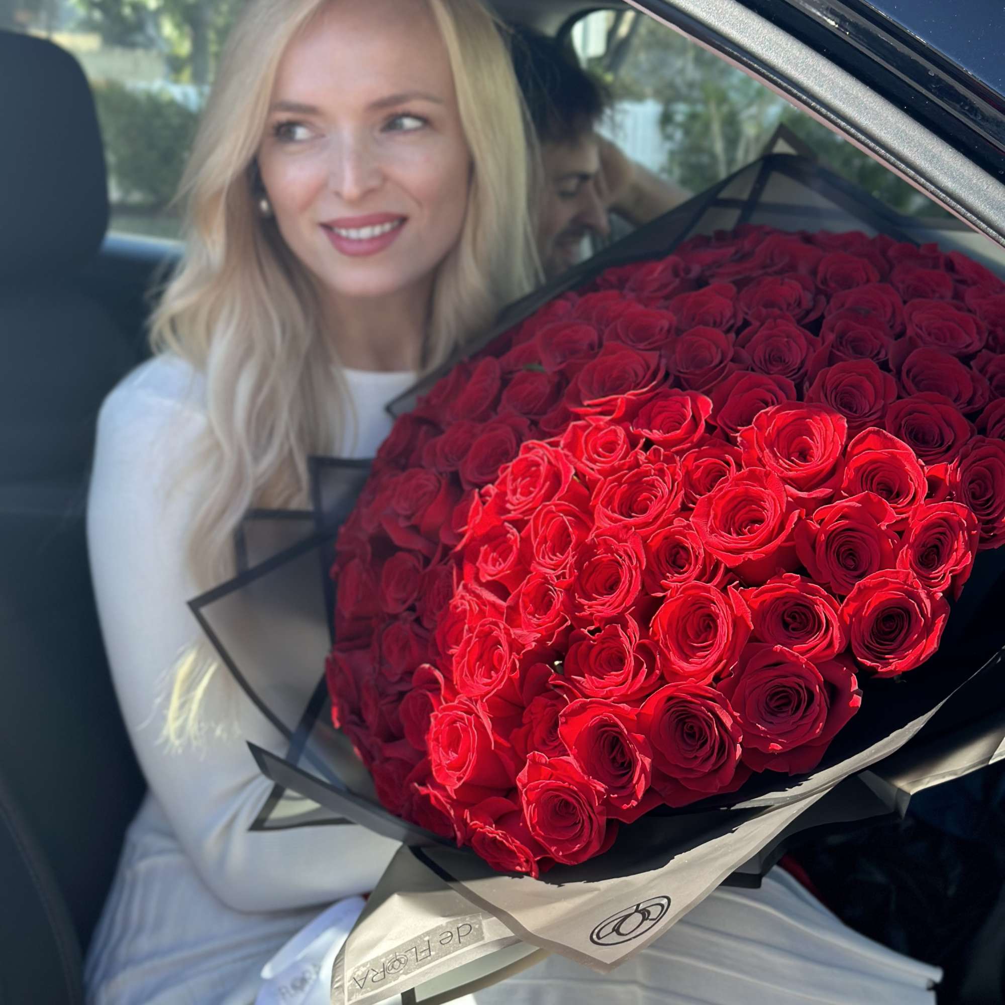 Large bouquet of tightly packed red roses wrapped in black paper, held by a woman in a car seat