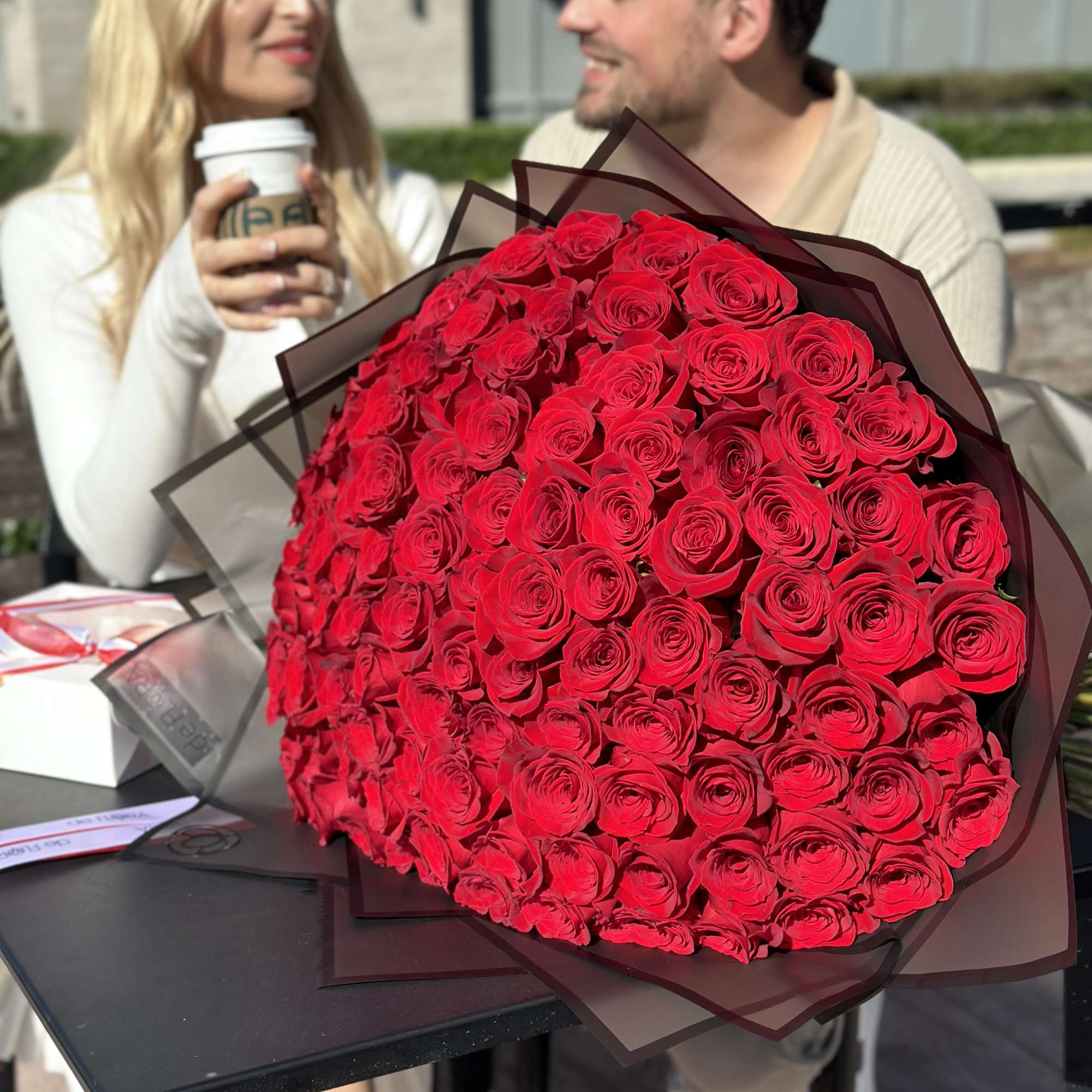 Large bouquet of red roses wrapped in dark paper on a table with a couple in the background