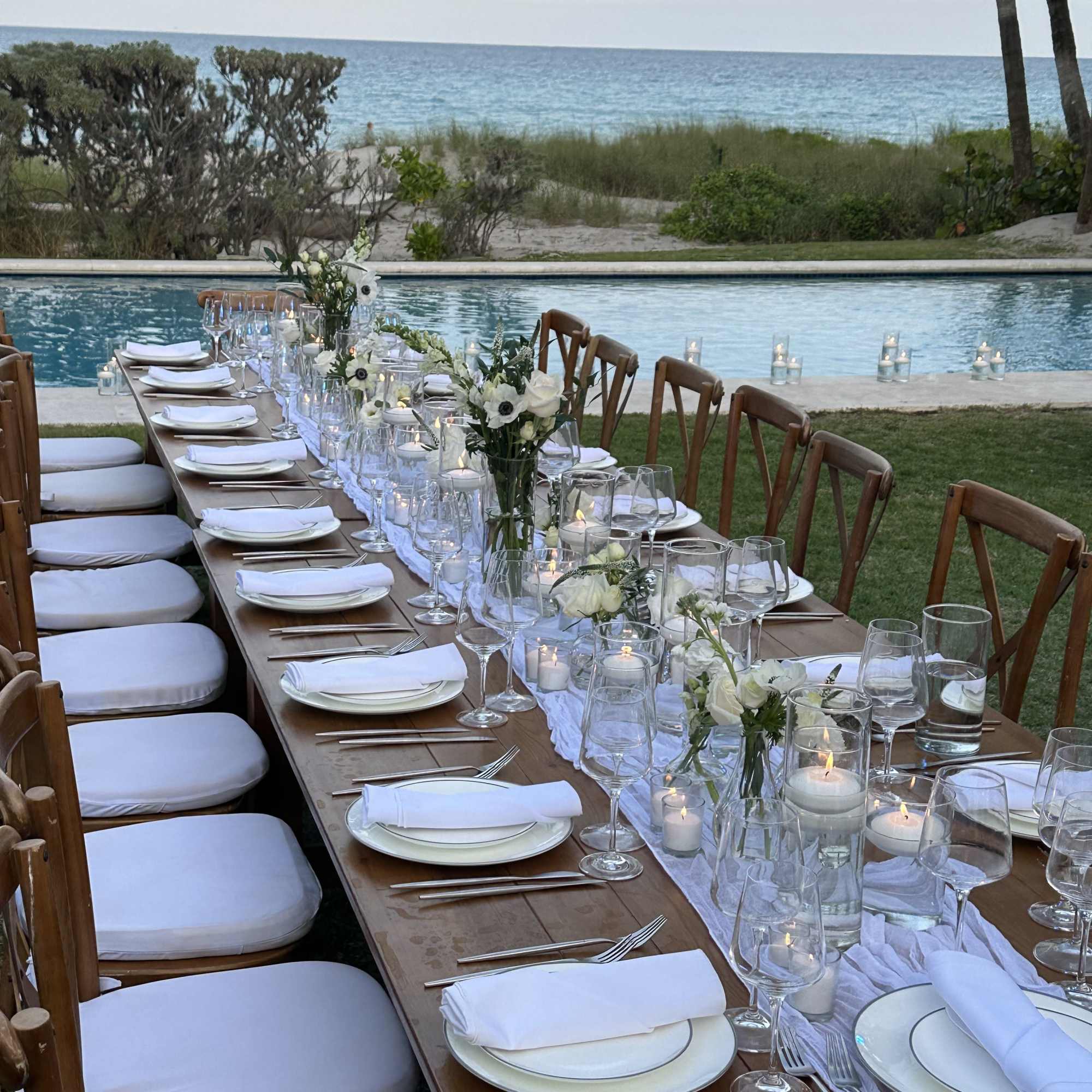 Long outdoor dining table with white floral centerpieces by a pool and ocean