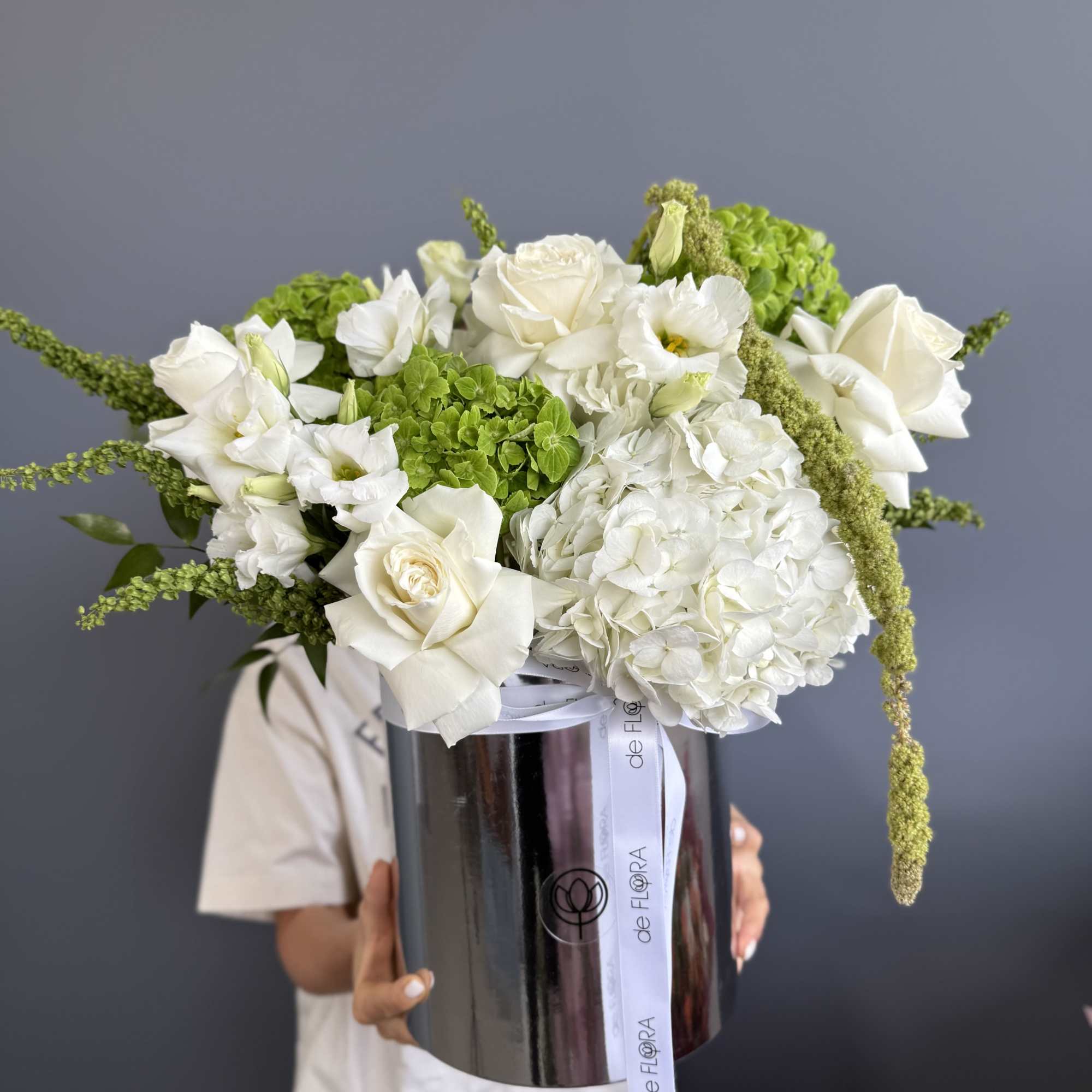 Large white and green floral arrangement of roses and hydrangeas in a tall metallic hatbox-style container