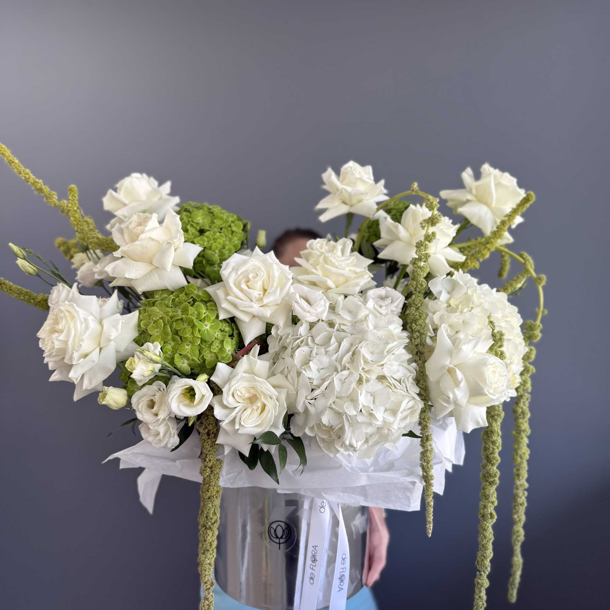 Tall white and green flower arrangement in a clear cylindrical box with trailing green accents