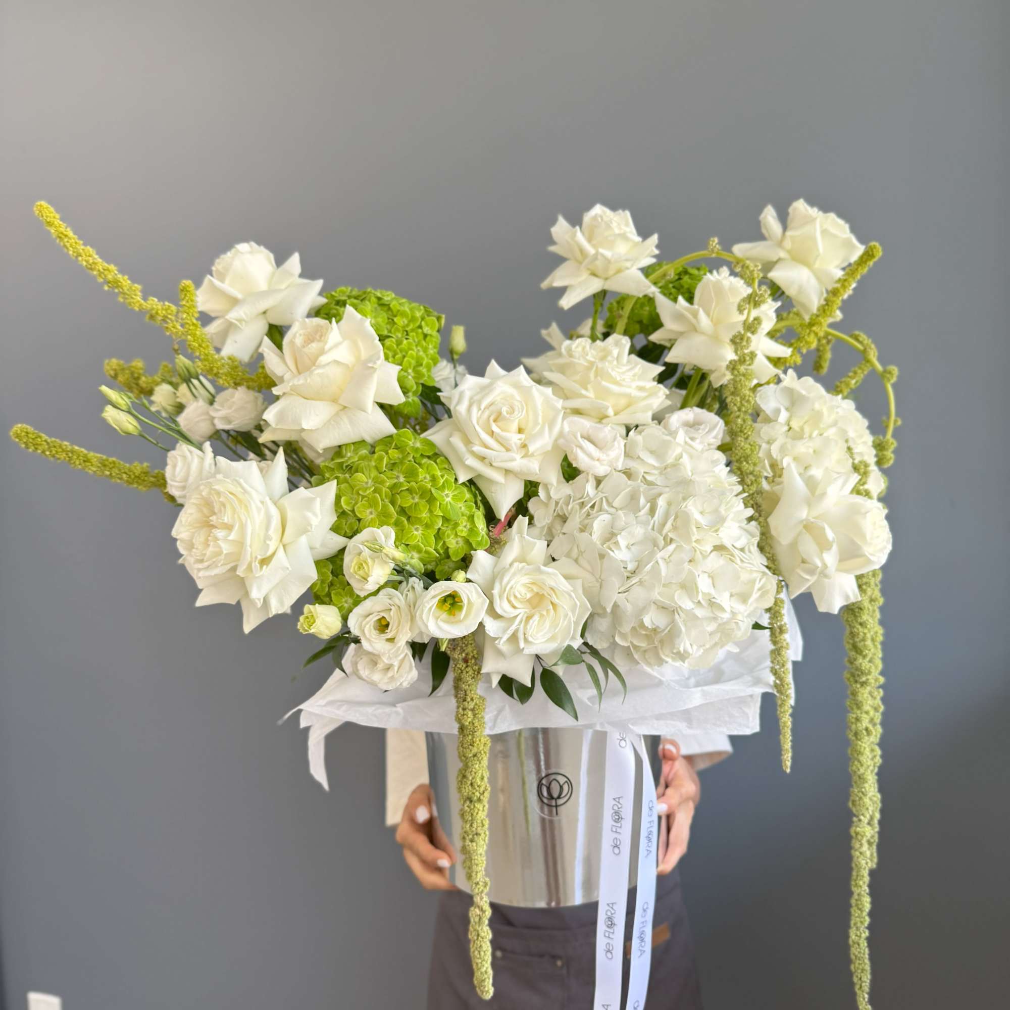 Tall arrangement of white roses and hydrangeas with green accents in a silver hatbox-style container.