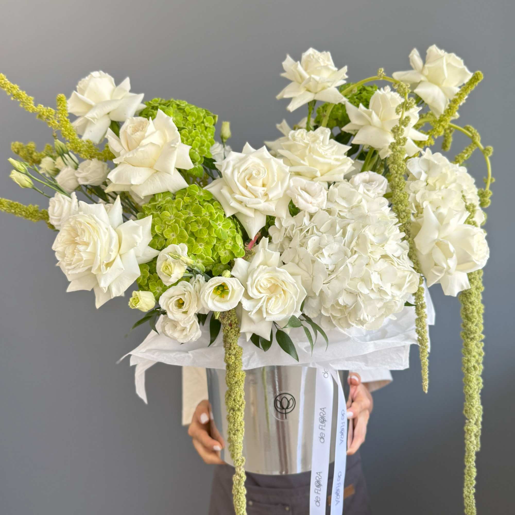 Large arrangement of white roses and hydrangeas with green accents in a silver hatbox container.