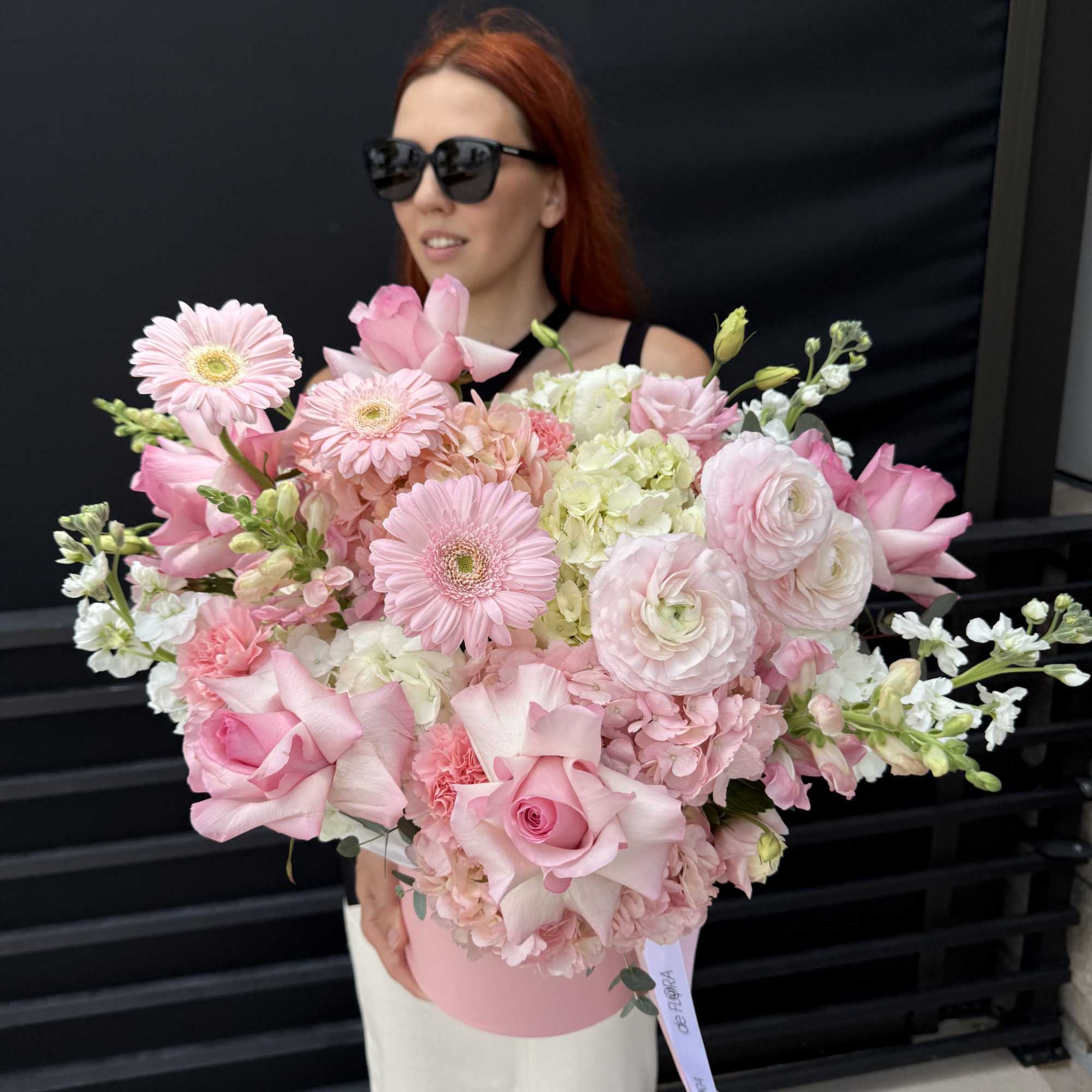 Large pastel pink flower arrangement in a round hat box held by a woman.