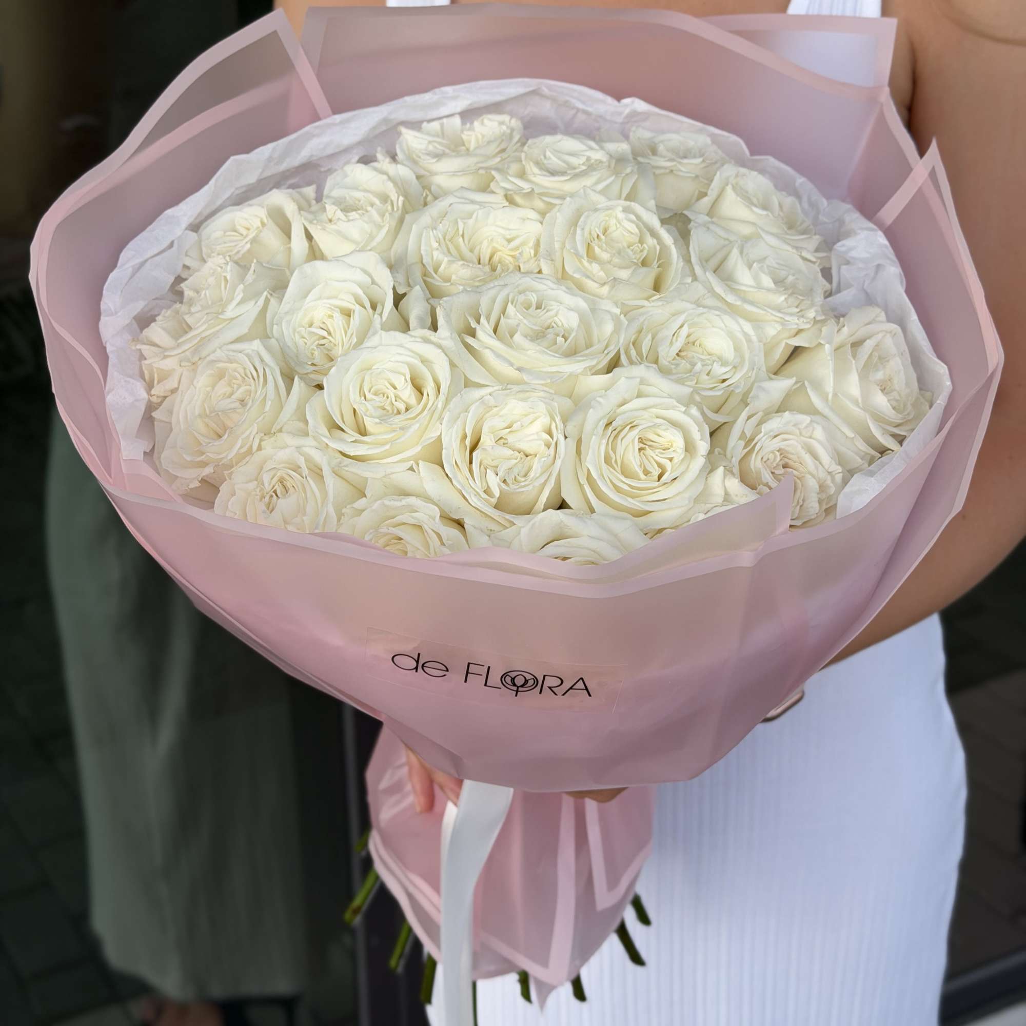 Large round bouquet of white roses wrapped in pale pink paper, held by a person in a white dress