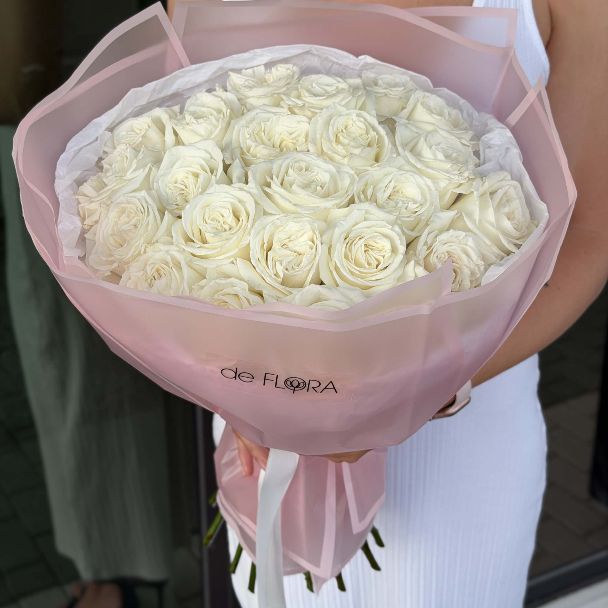 Large bouquet of white roses wrapped in pale pink translucent paper, held by a person in a white dress