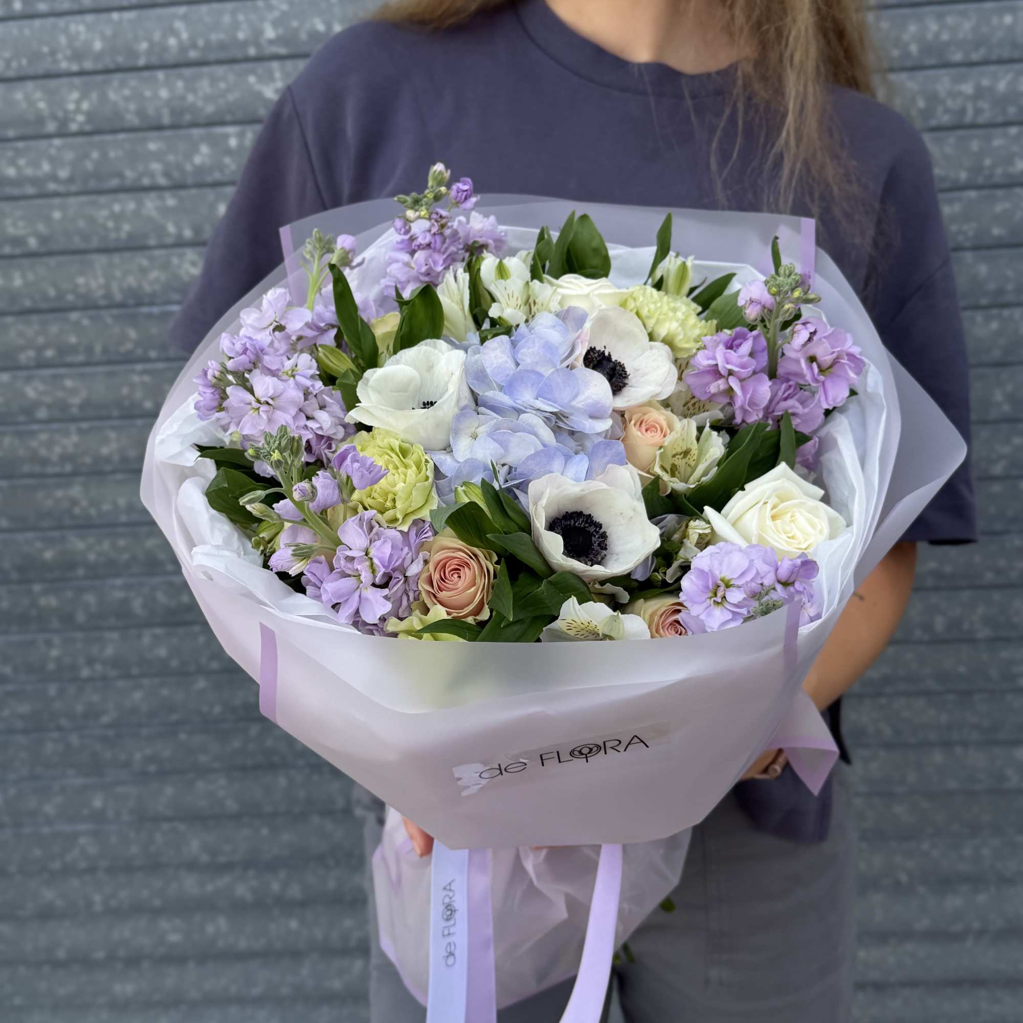 Pastel bouquet of white anemones, roses, and hydrangea wrapped in frosted paper, held by a person.
