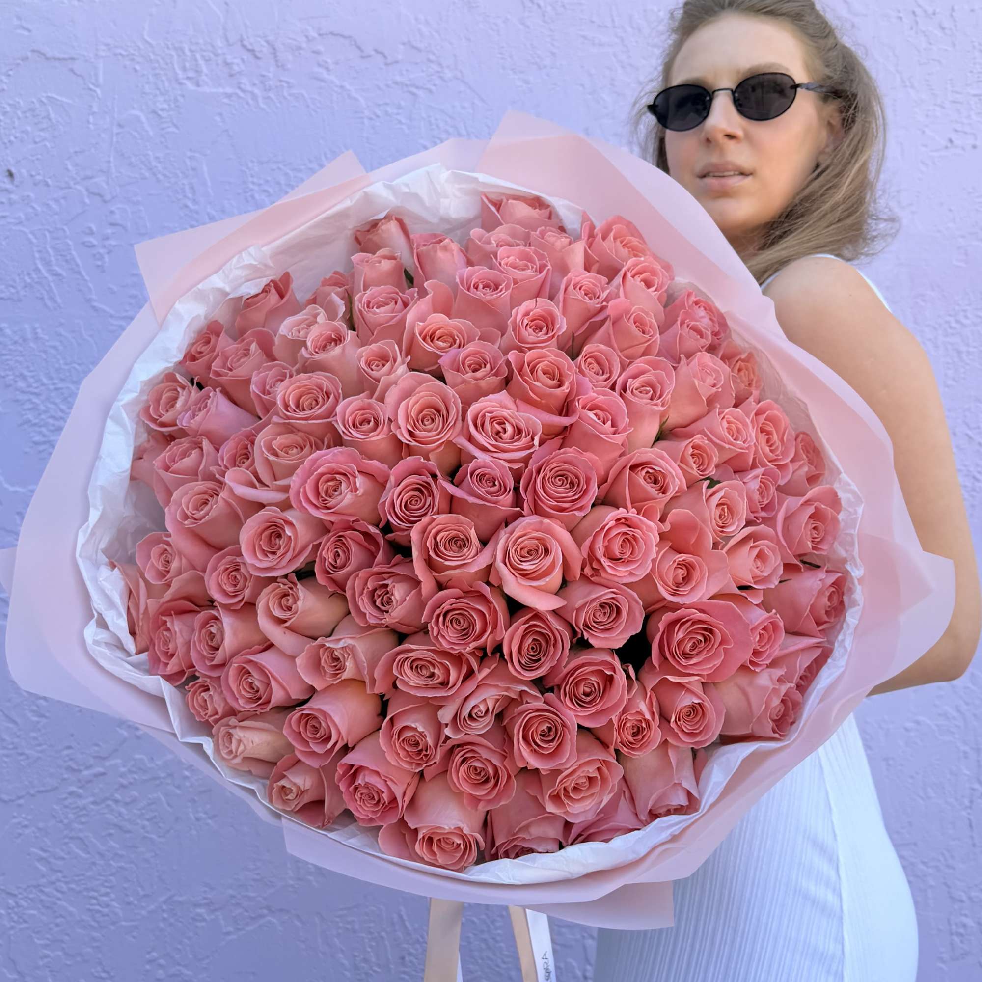 Large bouquet of tightly clustered pink roses wrapped in paper, held by a woman in sunglasses.