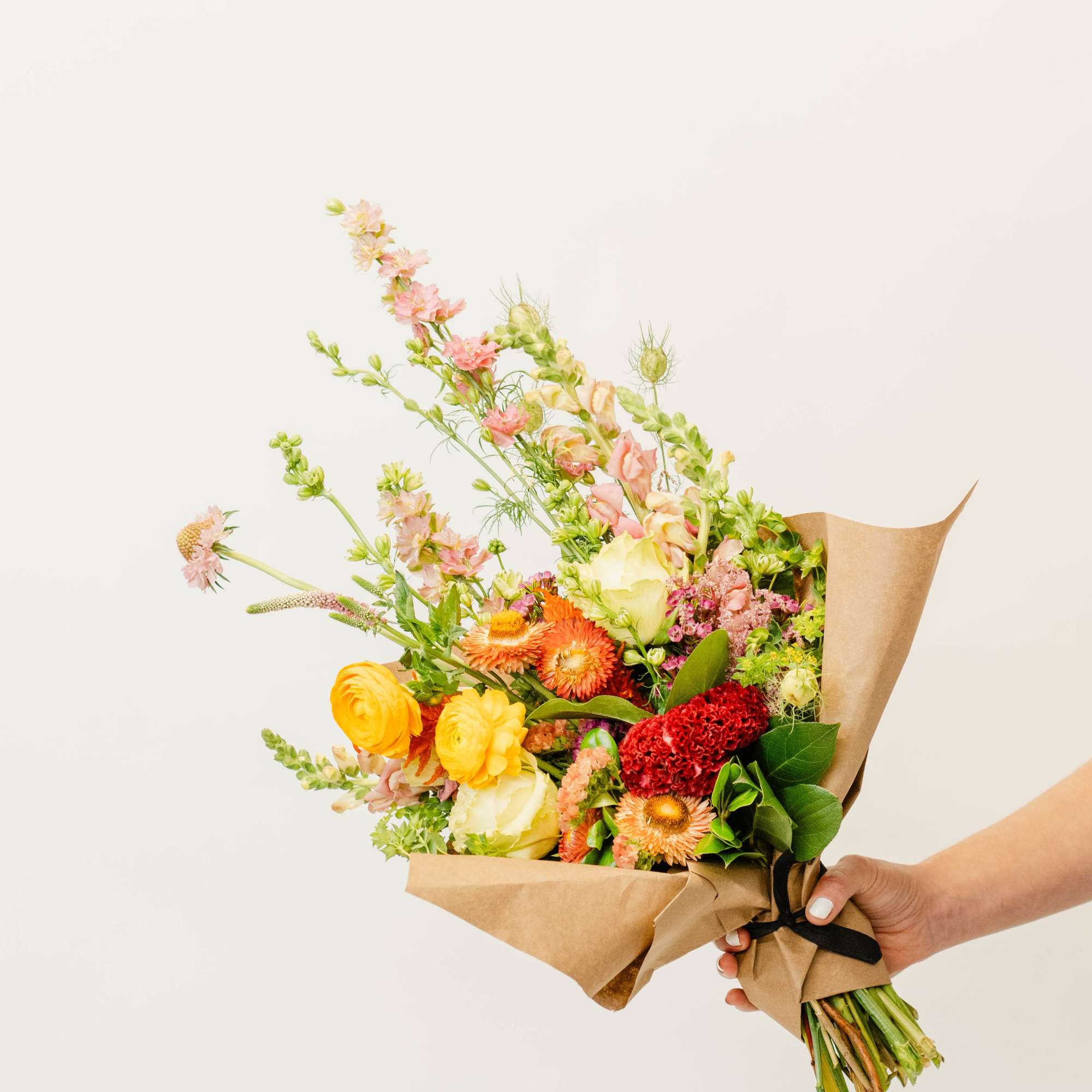 Handheld bouquet of mixed pink, yellow, orange, and white flowers wrapped in kraft paper