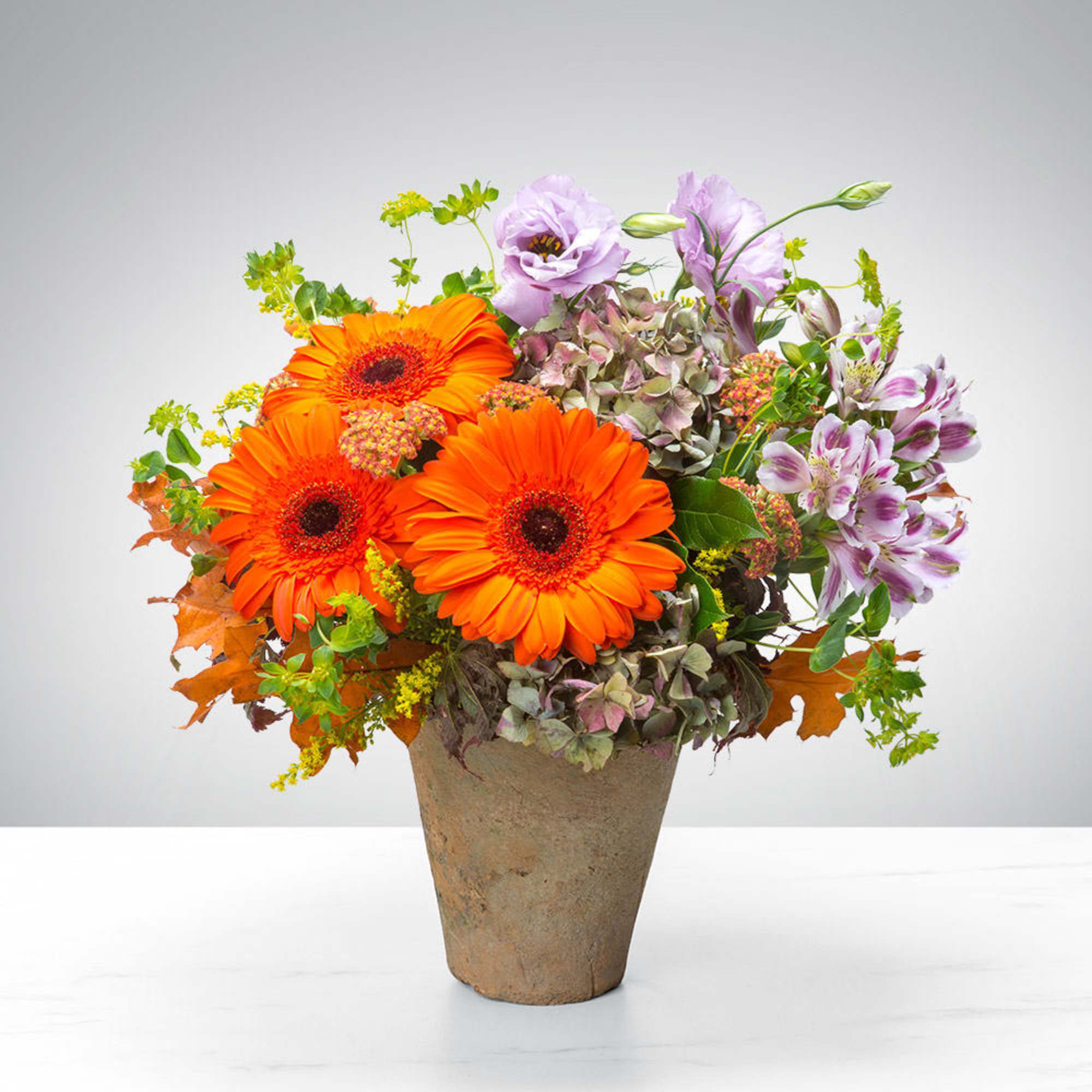 Orange gerbera daisies and lavender blooms in a rustic vase